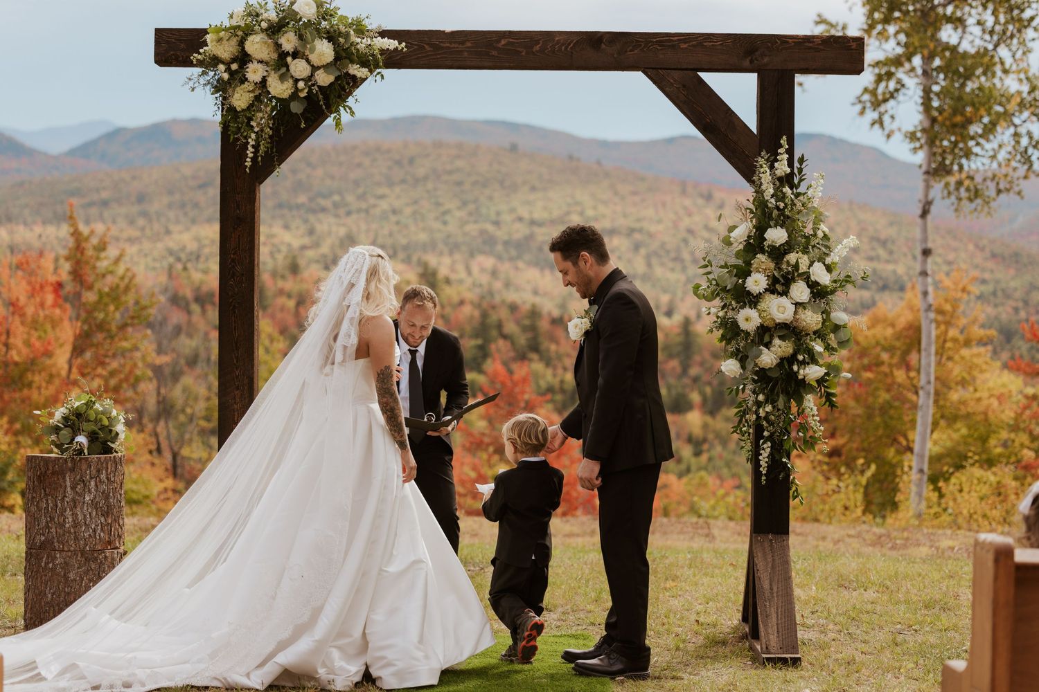 Outdoor fall wedding ceremony under rustic wooden arch with mountain foliage backdrop, bride groom and young ring bearer.