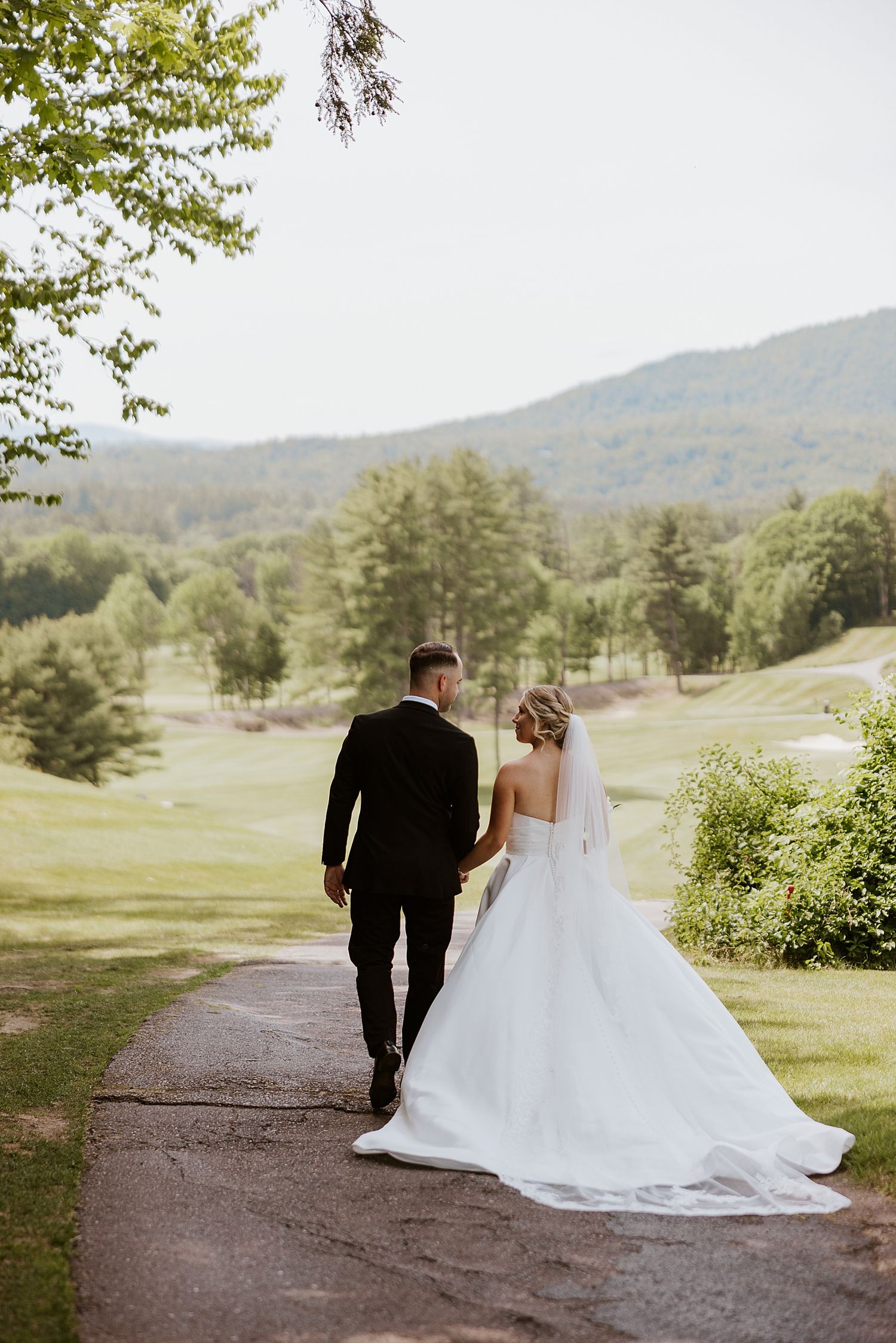 Bride and groom walking hand in hand on a path with lush green trees and rolling hills in the background.