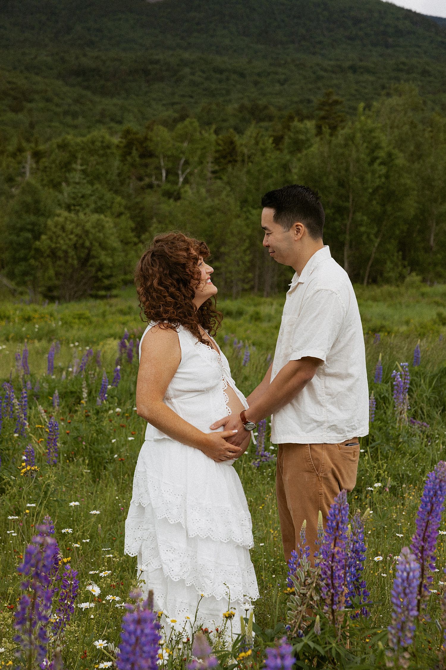 Pregnant couple holding hands in a wildflower field with purple lupines, mountains in background, maternity photo session.