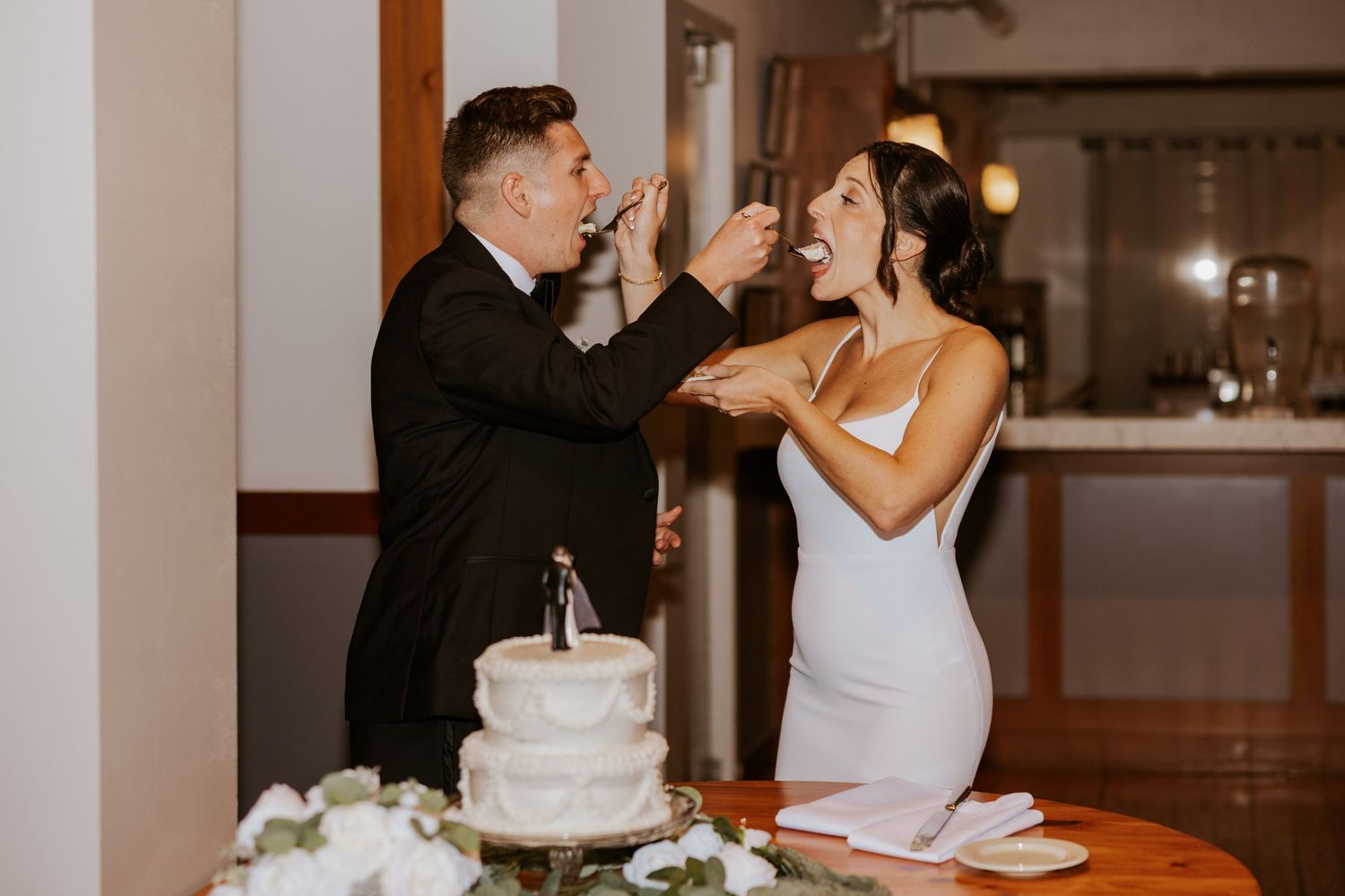 Bride and groom playfully feeding each other wedding cake at reception.