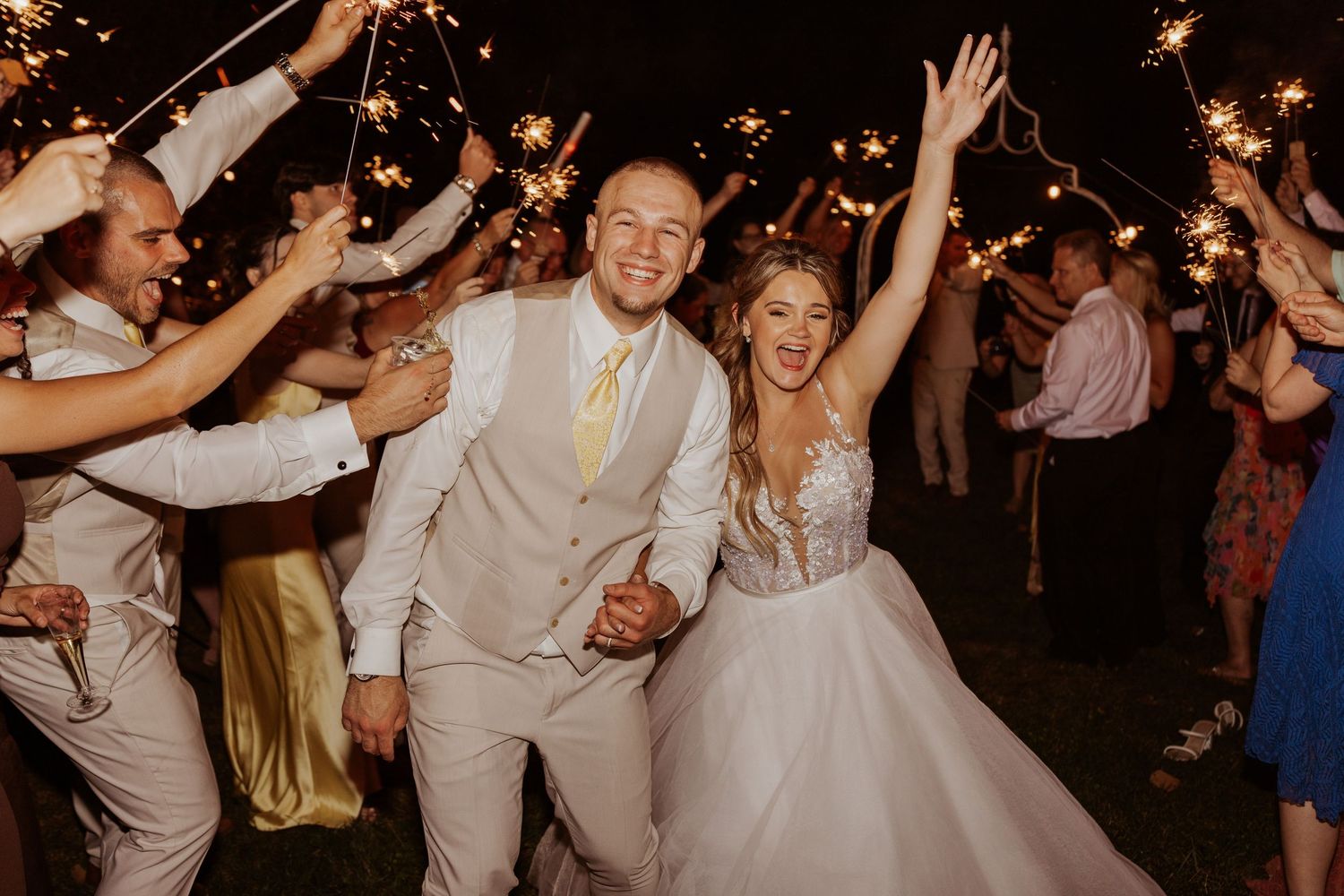Joyful bride and groom walking through sparkler sendoff surrounded by celebrating wedding guests.