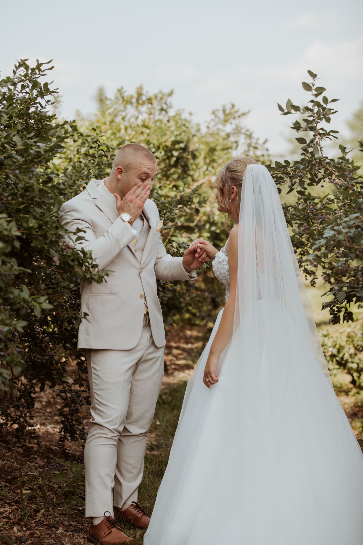 Emotional groom covers his mouth in awe seeing his bride in her white wedding gown outdoors.