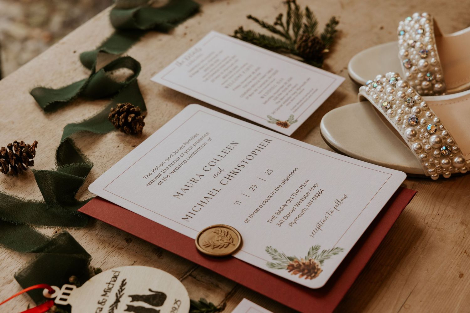 Elegant winter wedding invitation suite with gold wax seal, pine cones, and green ribbon on wood.