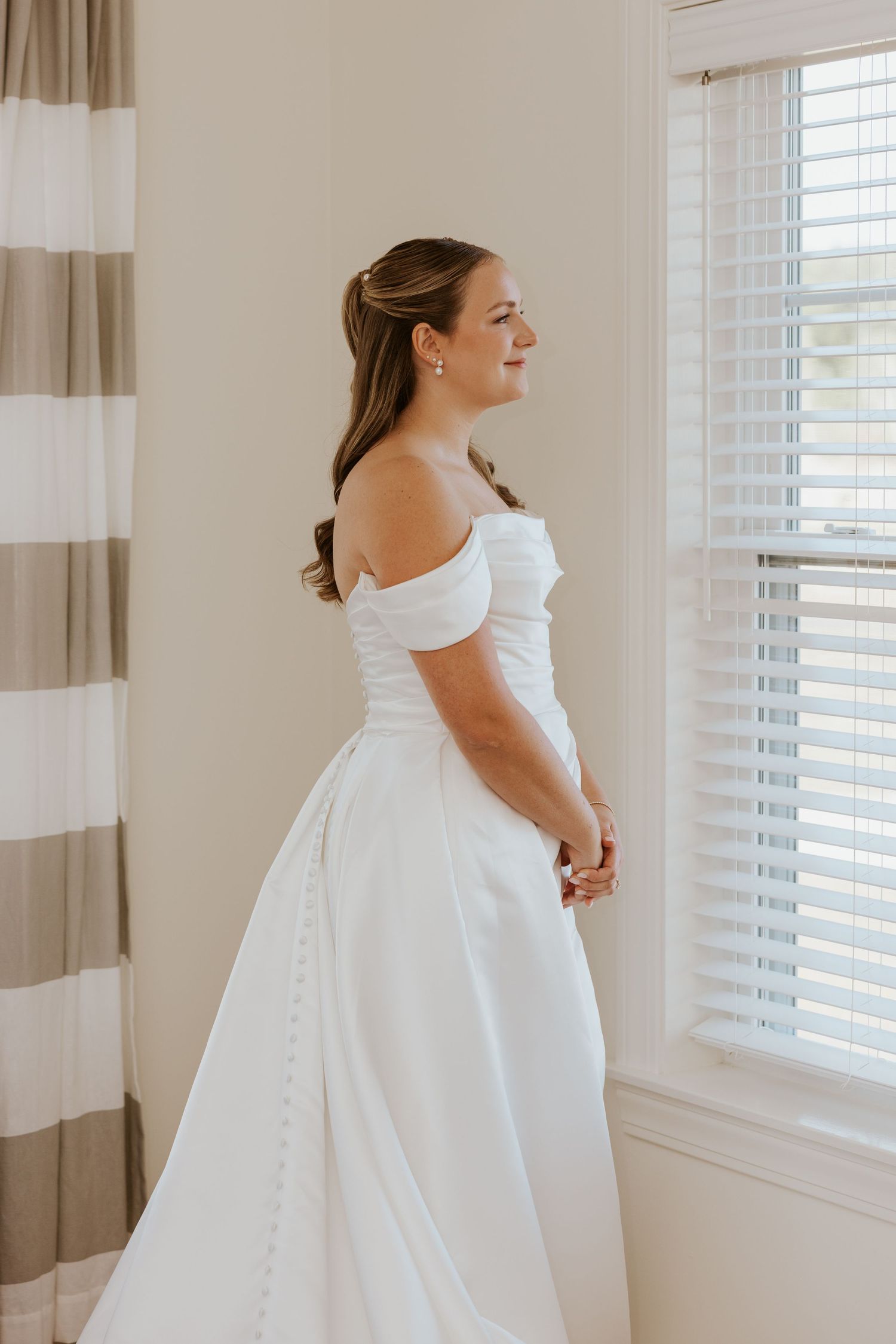 Elegant bride in white off-shoulder satin gown with cathedral train gazing out a sunlit window.