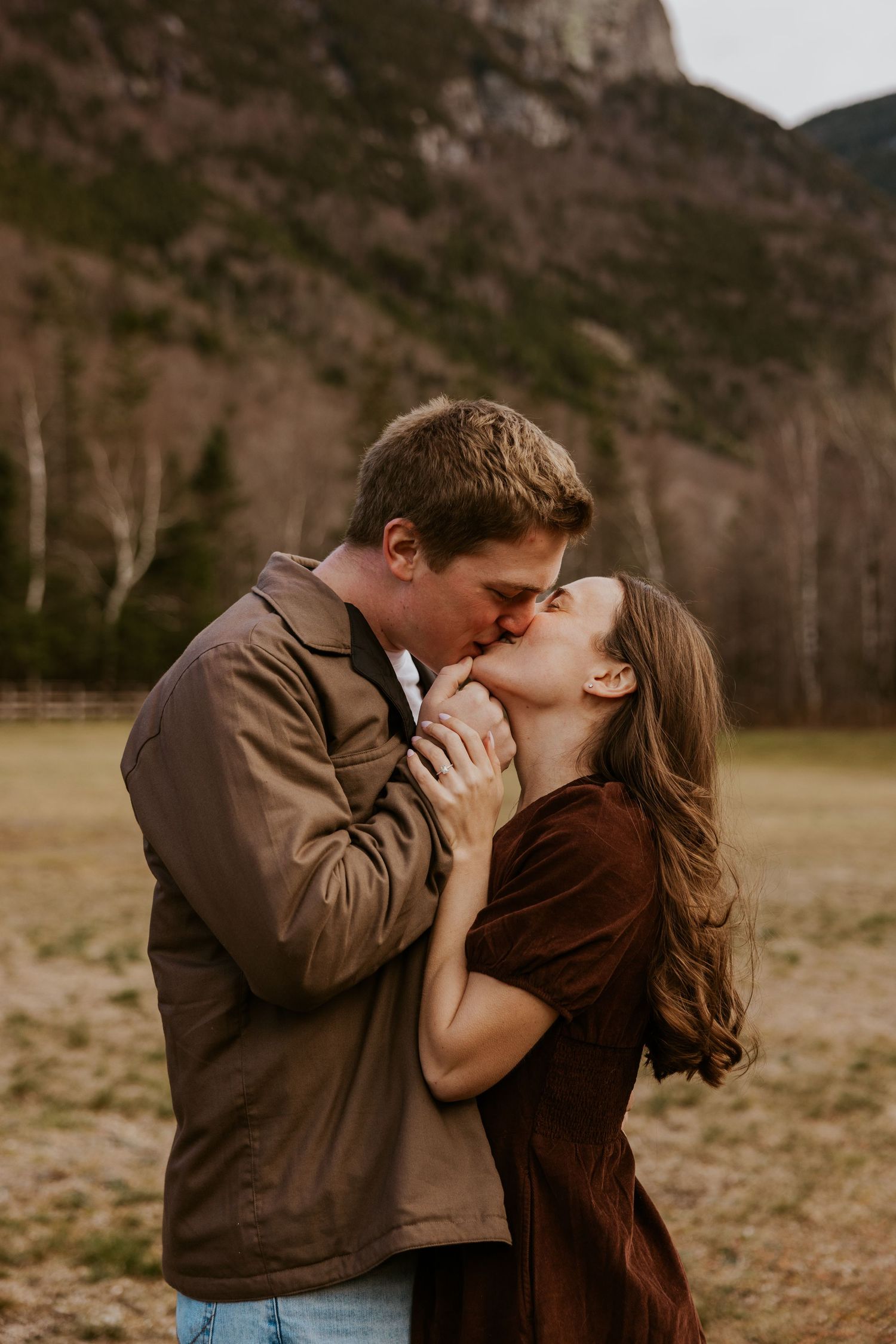 Couple sharing a passionate kiss in a mountain meadow with rocky cliffs and pine trees in the background.