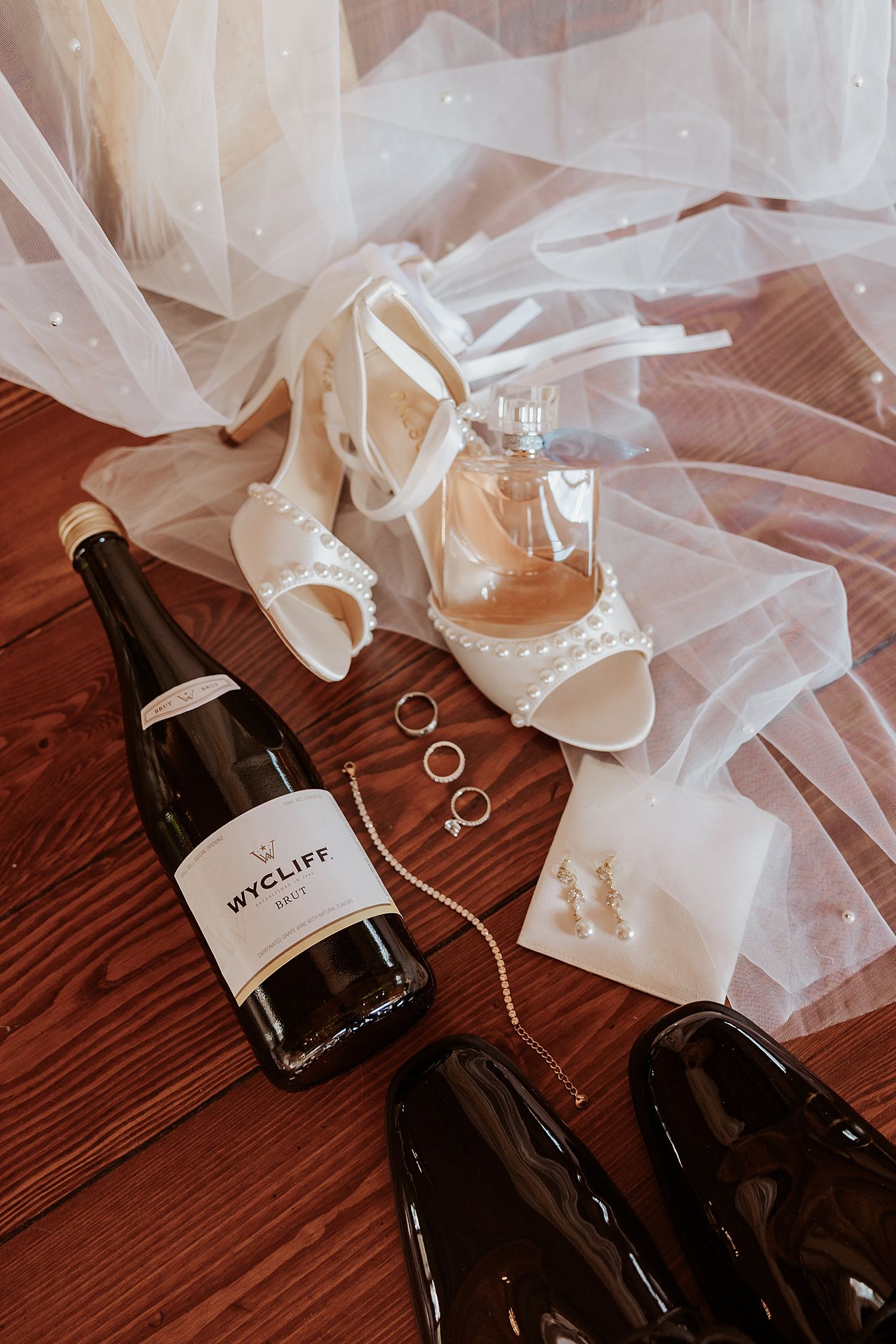 Wedding flat lay with Wycliff champagne, pearl heels, rings, earrings, and bridal veil on rustic wood floor in New Hampshire