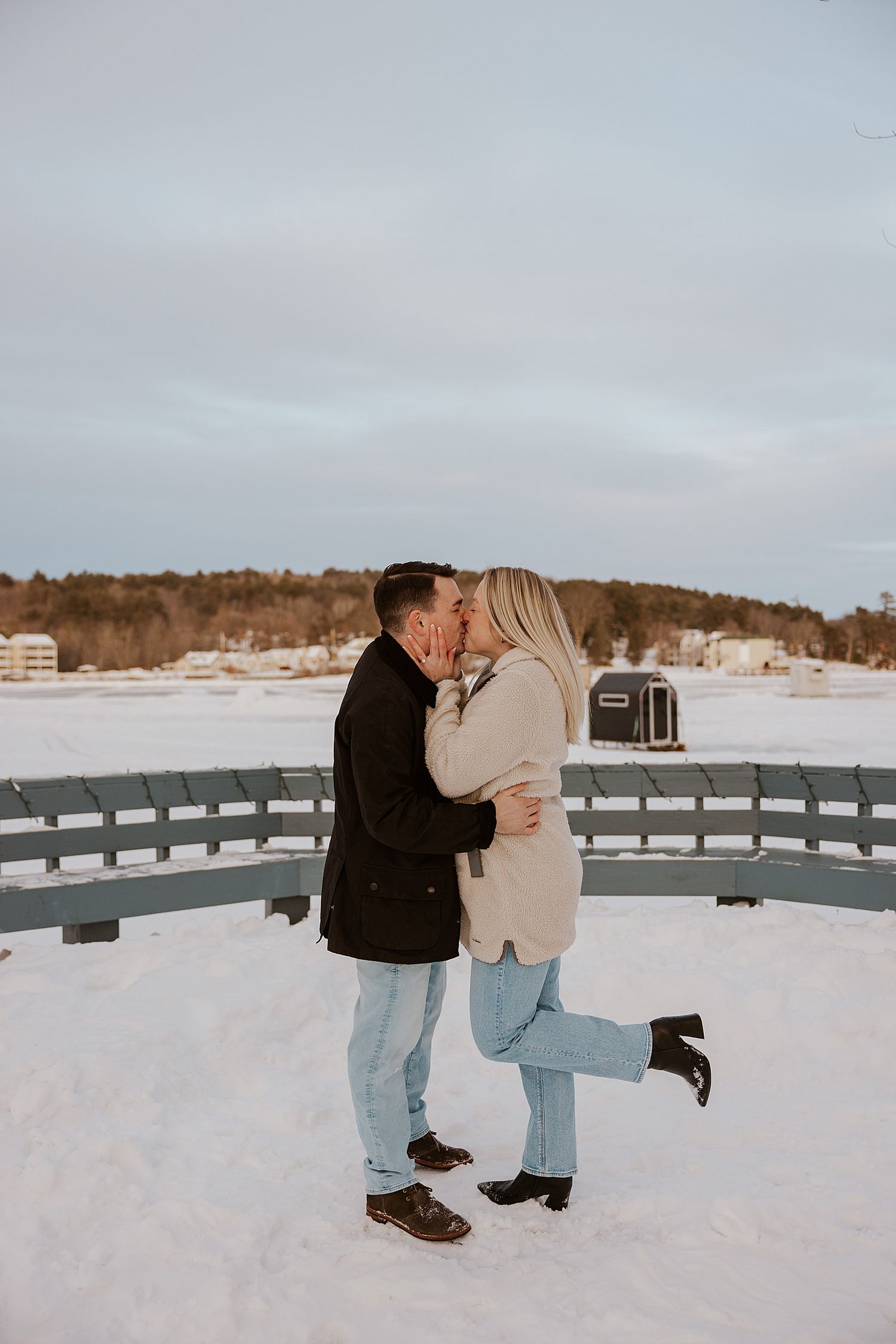 Couple sharing a romantic kiss on a snowy bridge in winter, dressed in casual coats and jeans.