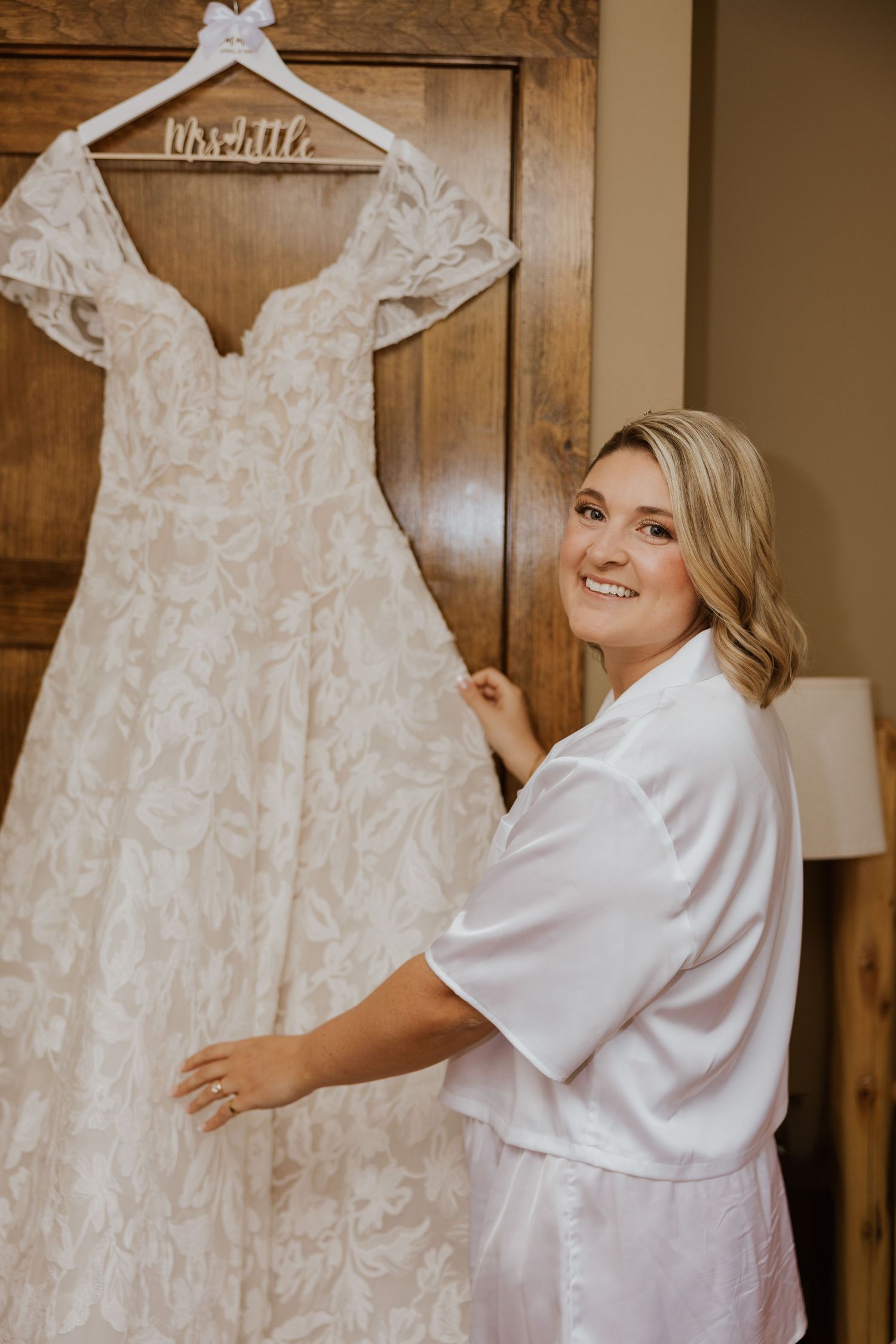 Bride in white satin robe smiling beside her lace wedding gown hanging on a wooden door.