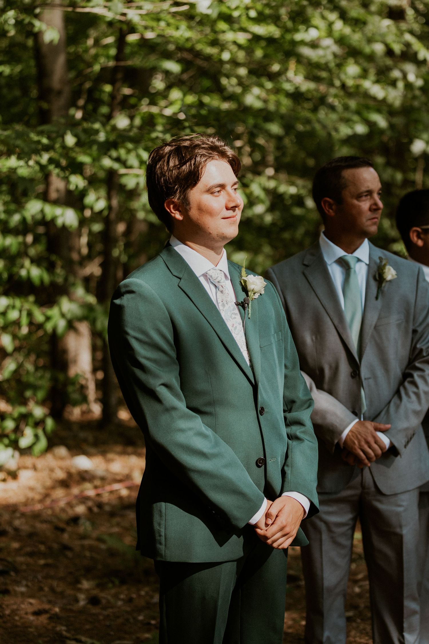 Wedding party members stand in formation wearing green and grey suits in a woodland setting.
