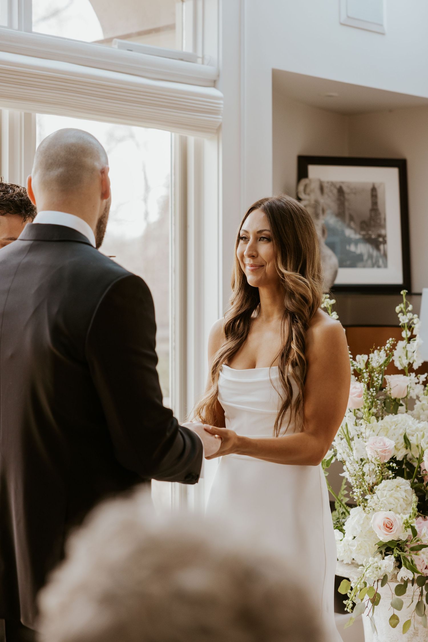 A romantic first look moment between bride and groom in a sunlit room with white floral arrangements.