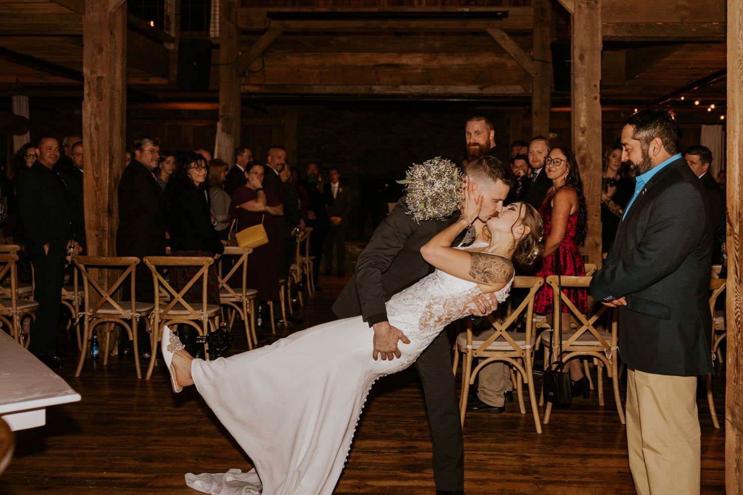 Groom dips bride on dance floor during first dance at rustic barn wedding reception with guests watching.
