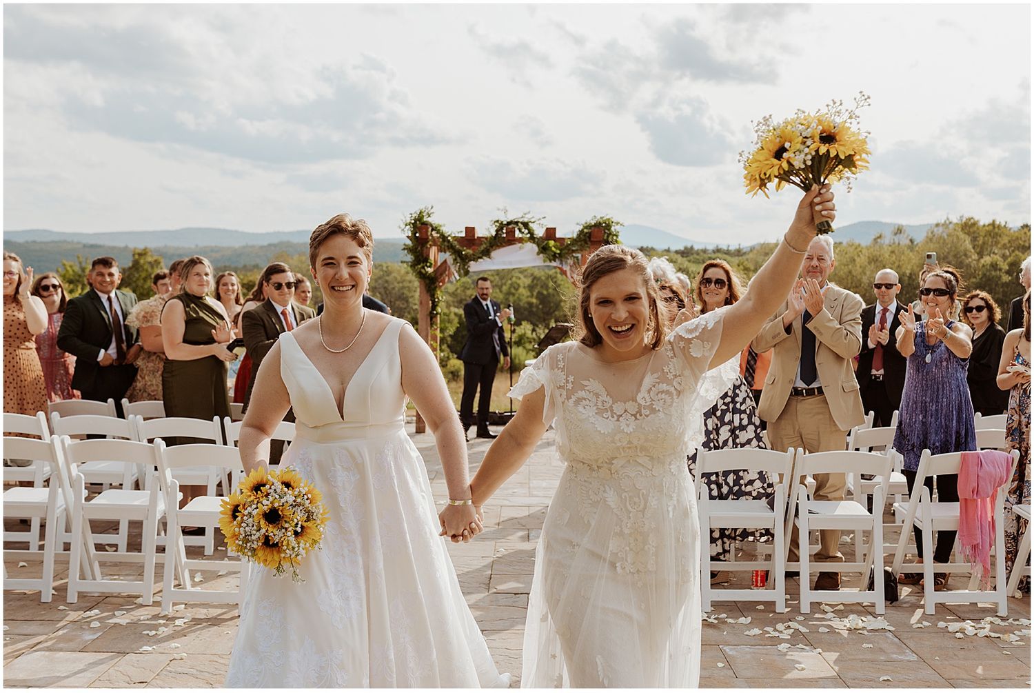 Wedding guests celebrate during an outdoor ceremony with white chairs and yellow sunflower bouquets.