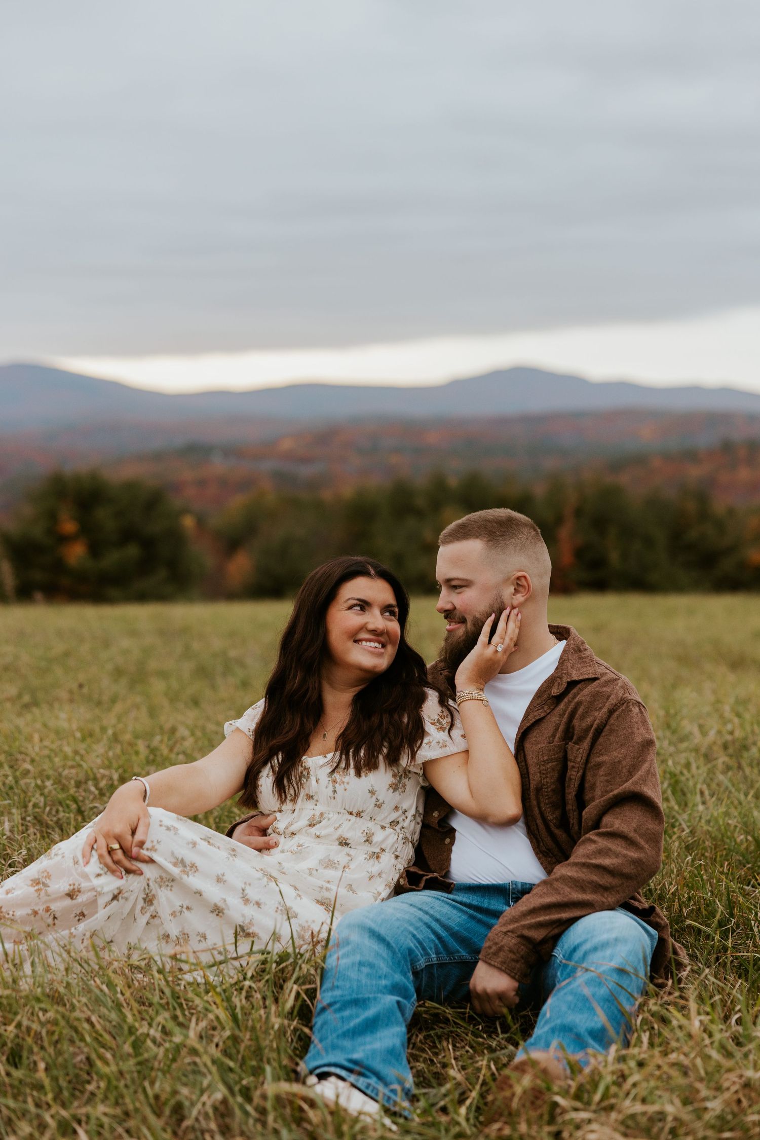 Engaged couple sitting in a grassy field, gazing at each other with colorful fall mountains behind them.