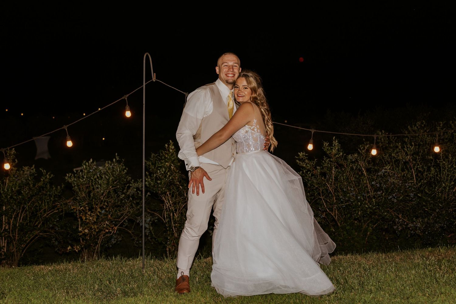 Bride and groom posing together under string lights at night during outdoor wedding reception.