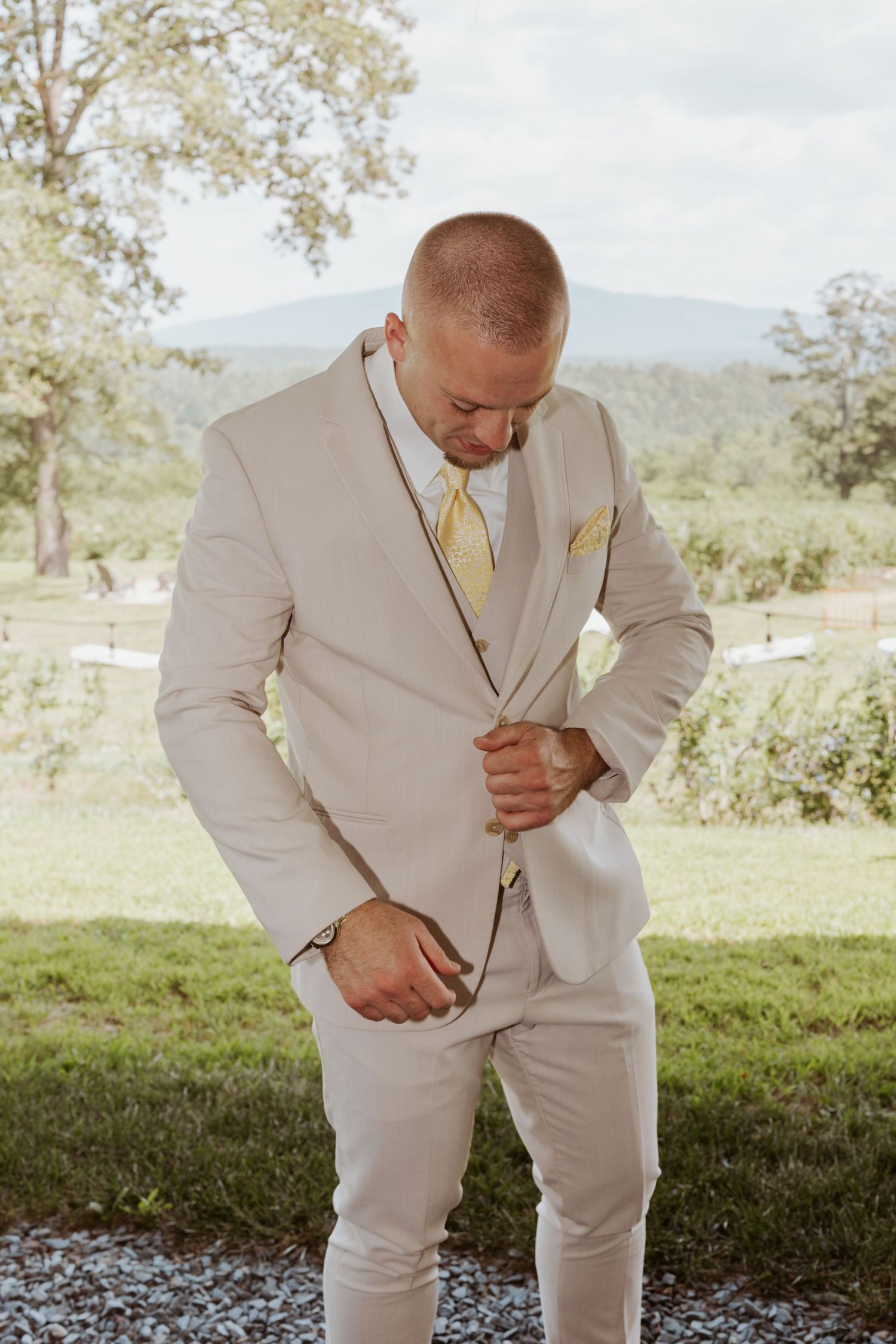 Dapper groom in a beige suit with yellow tie adjusting his jacket outdoors with mountain views.