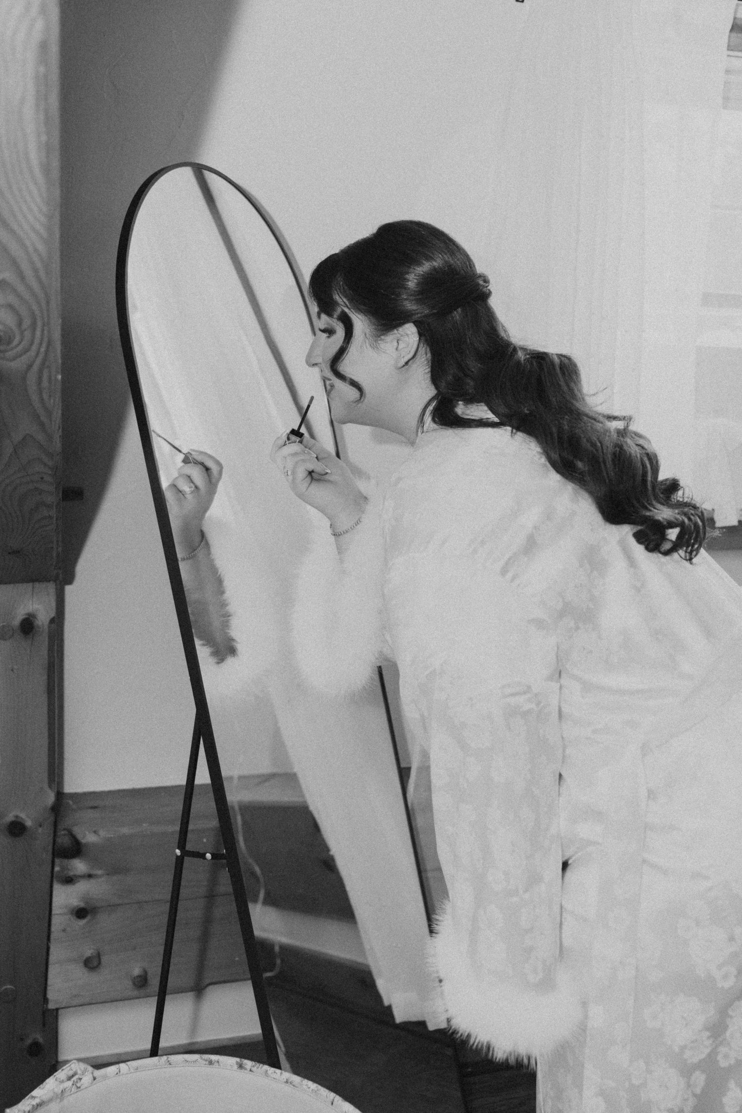 Black and white photo of bride in lace gown and fur wrap looking into a standing mirror.