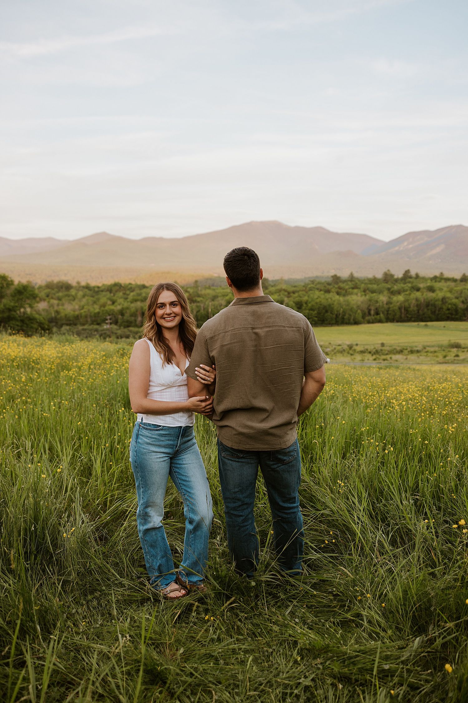 Couple holding hands in a lush green meadow with mountain views and wildflowers during golden hour.