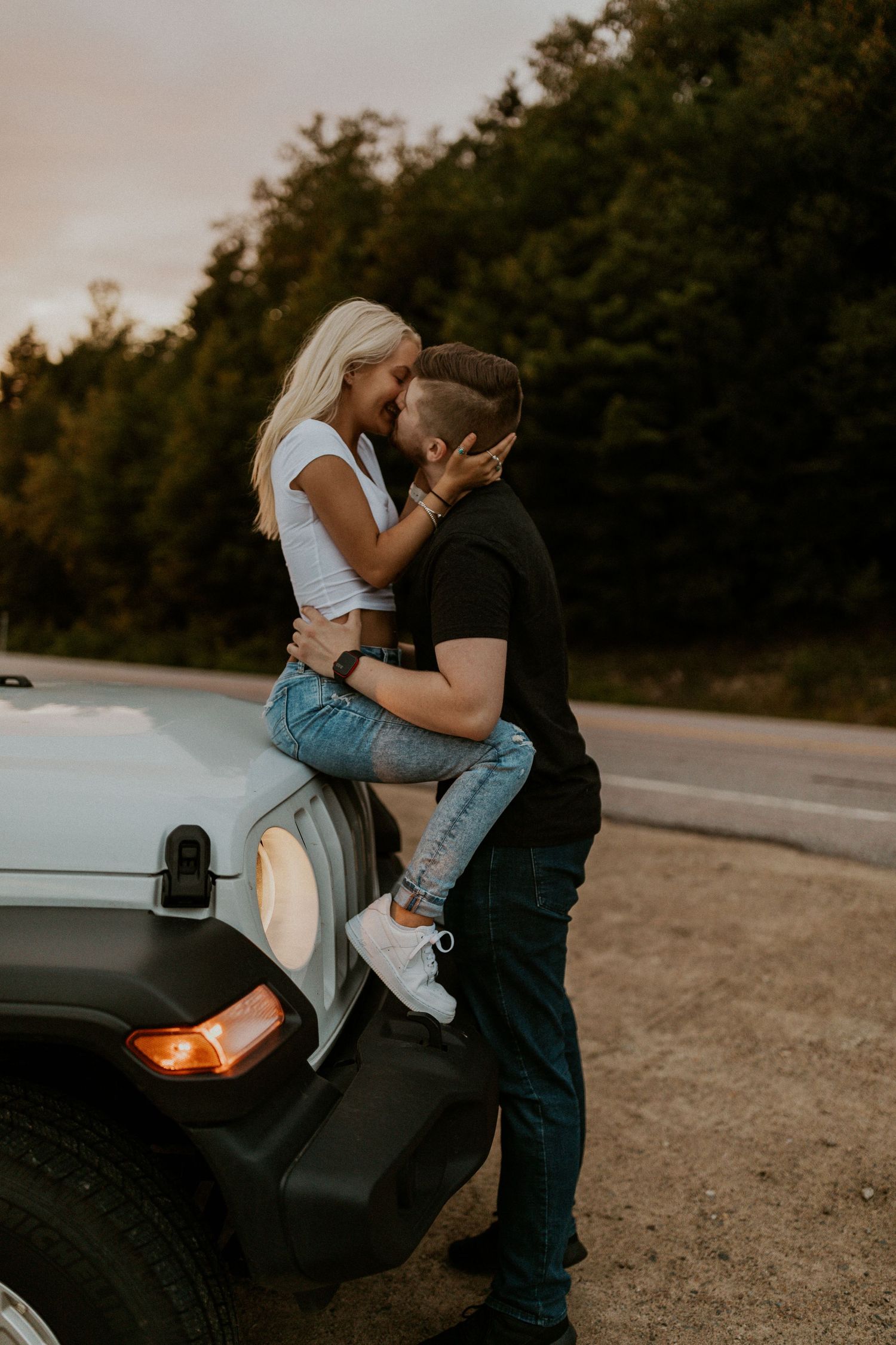 Paige is laying across the hood of her jeep as Justin stands beside her and ksises her at sunset in the White Mountains