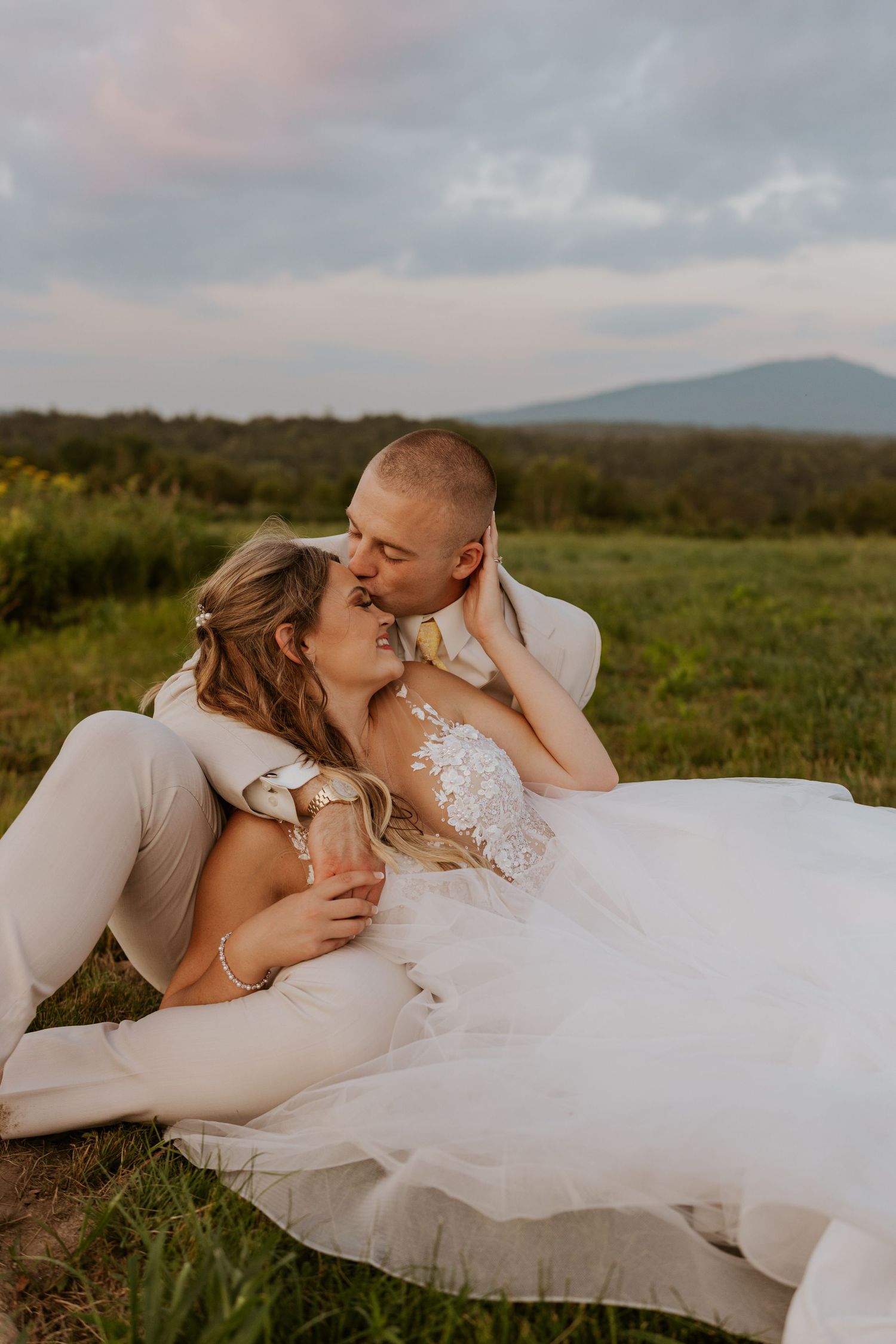Bride and groom sharing a kiss while sitting in a lush green field with mountain views.