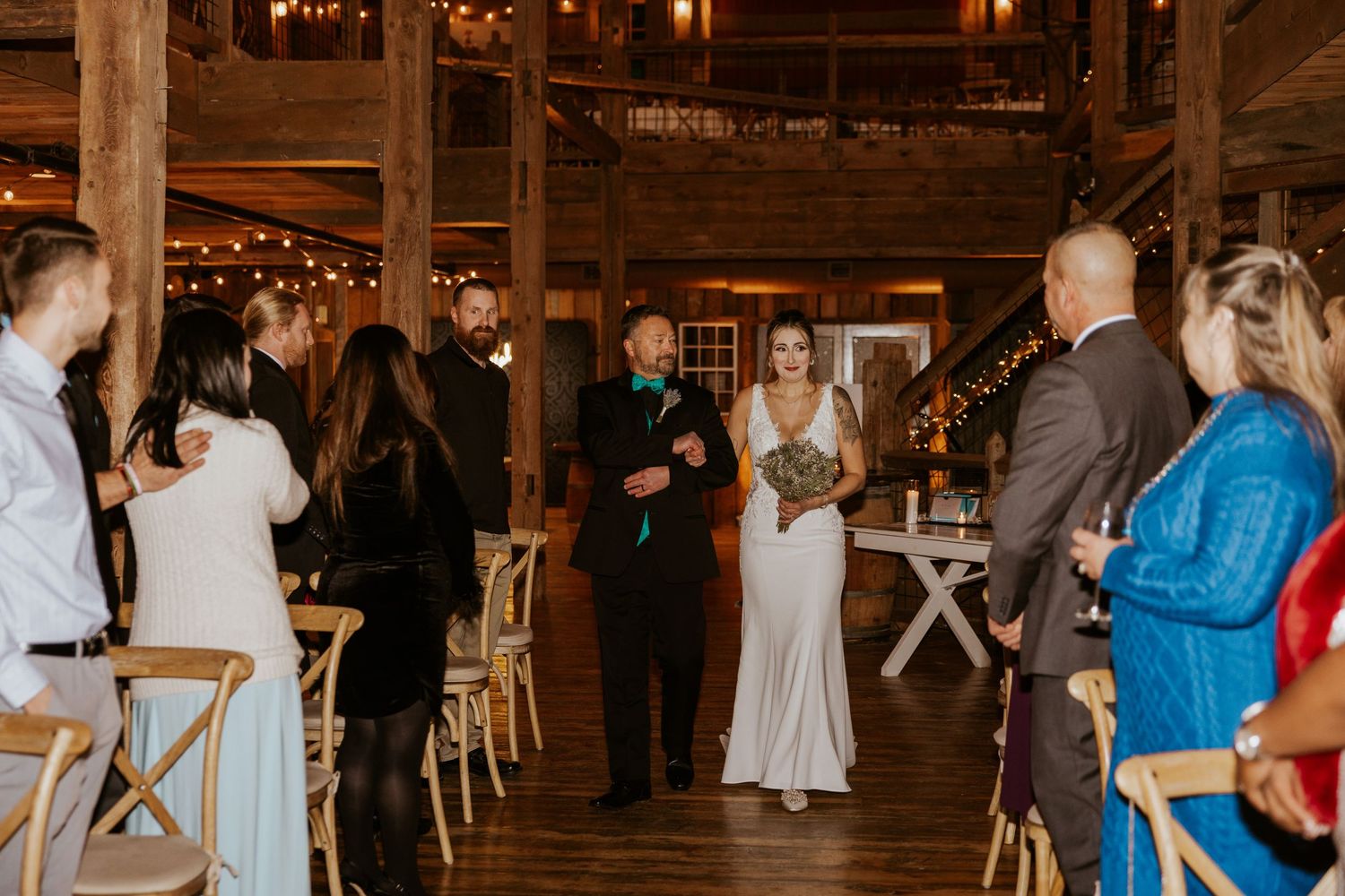 Bride and groom walk down the aisle in a warmly lit rustic barn wedding ceremony surrounded by guests.