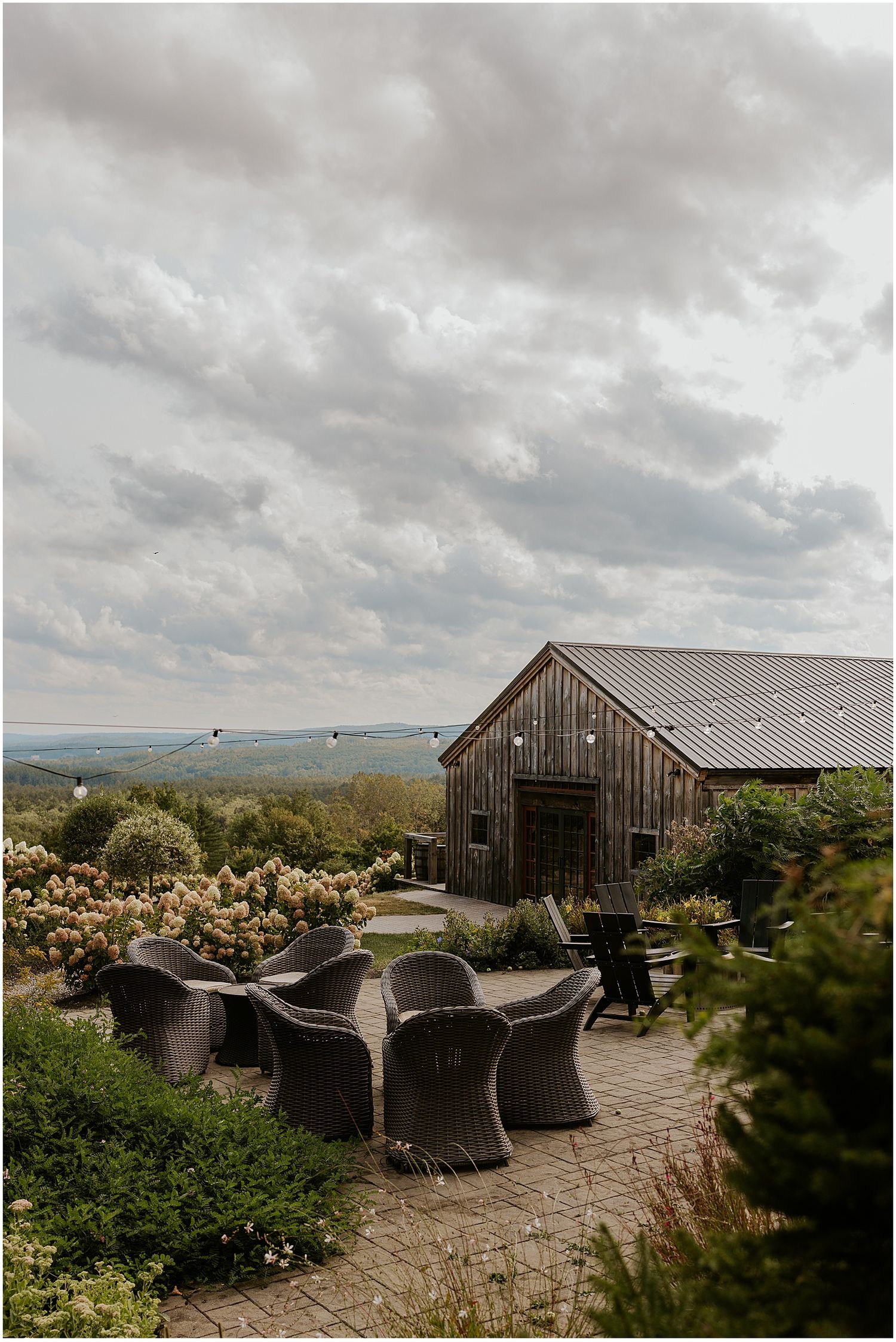 Rustic wooden barn with outdoor wicker seating area on stone patio overlooking scenic mountain landscape under cloudy skies.
