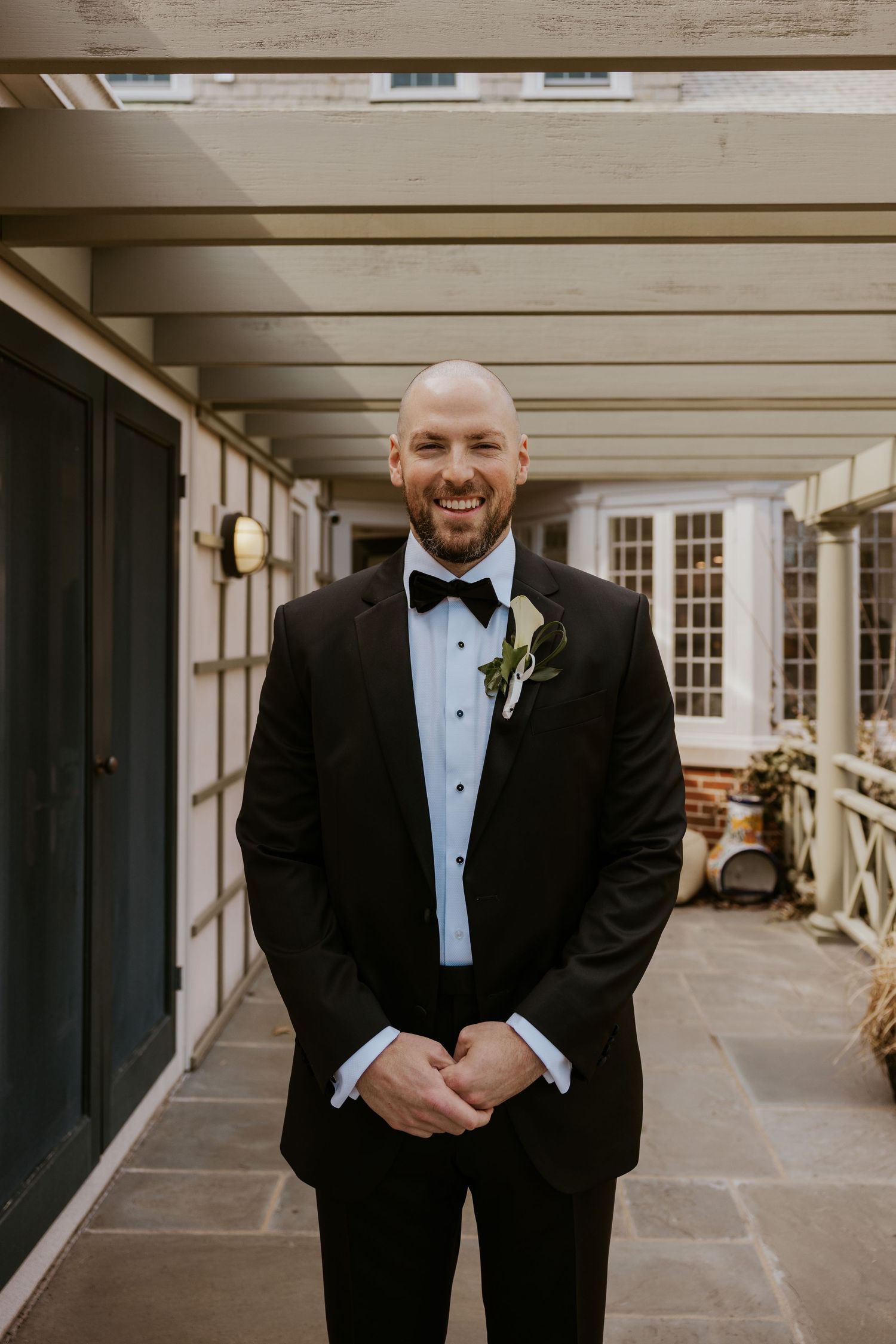 Groom in classic black tuxedo poses in elegant covered walkway with white columns and natural lighting.