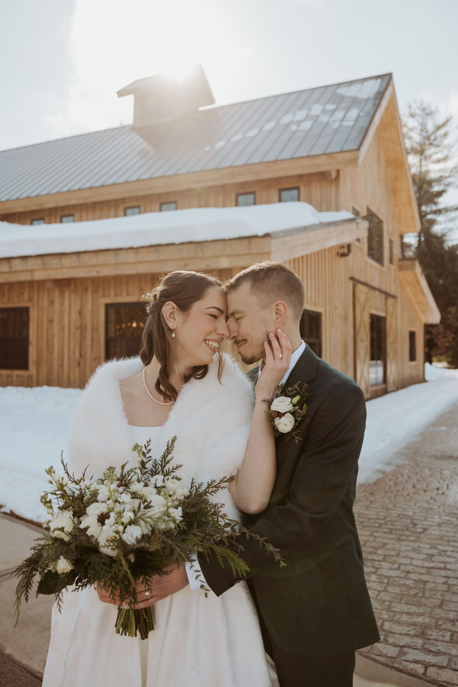 Winter wedding couple shares intimate moments outside rustic wooden barn venue in snowy setting.
