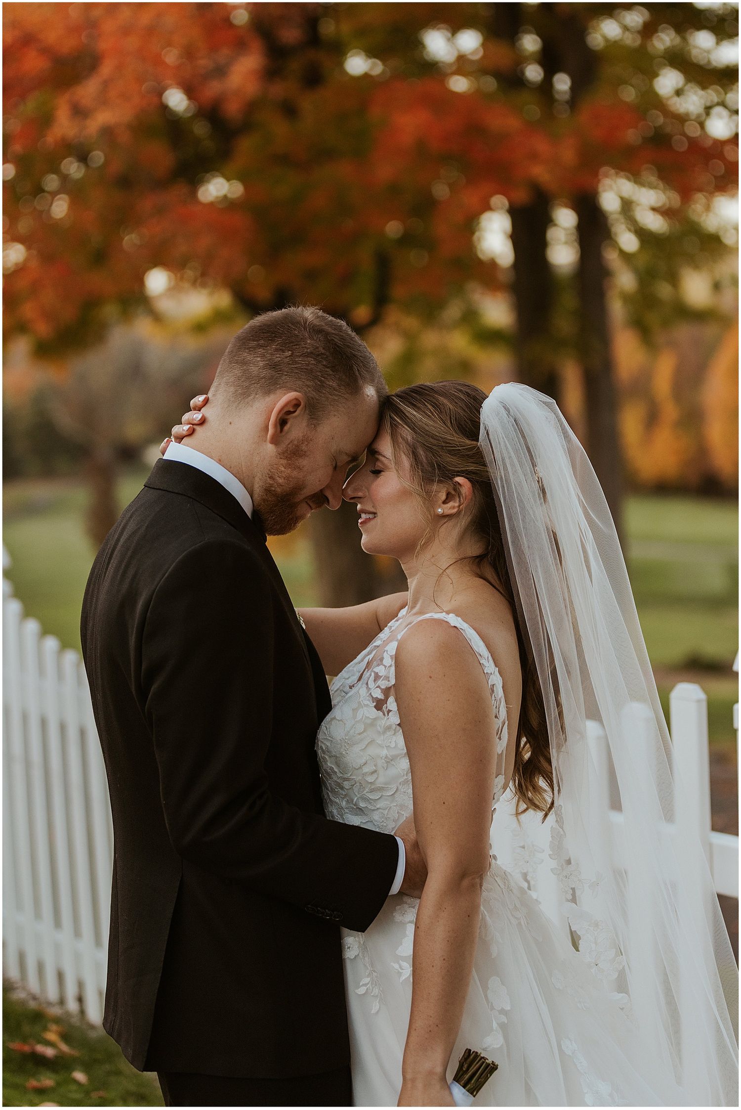 Romantic wedding portrait of couple embracing against vibrant autumn trees and white picket fence.