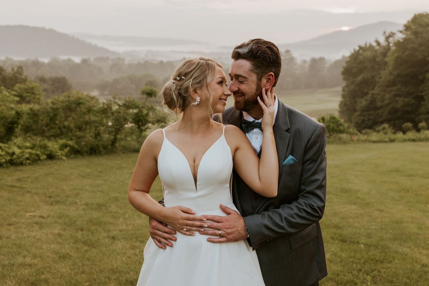 Bride and groom share a romantic moment in a lush green field with misty mountain views behind them.