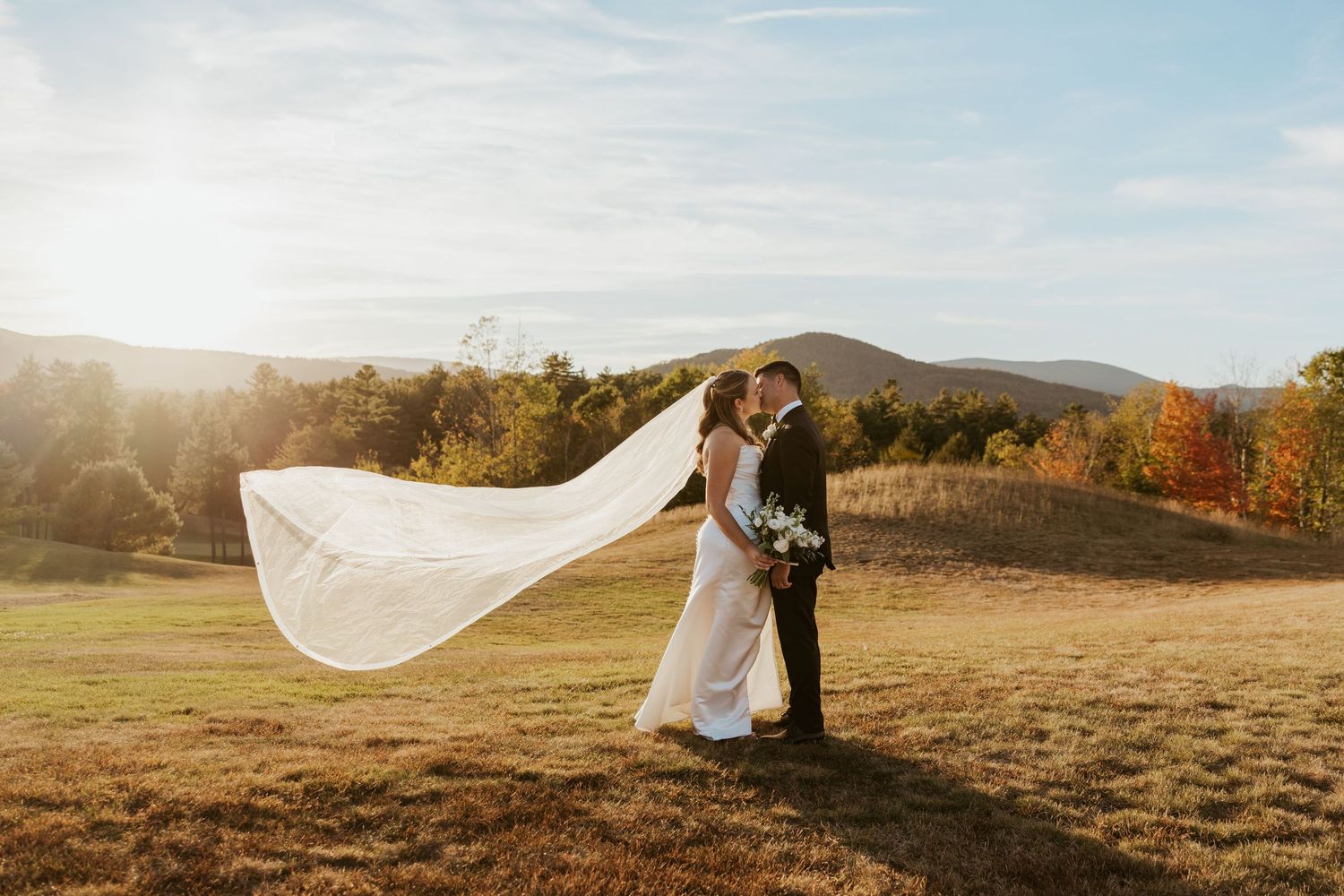 Bride and groom kiss in golden field with veil flowing in wind and autumn mountain backdrop.