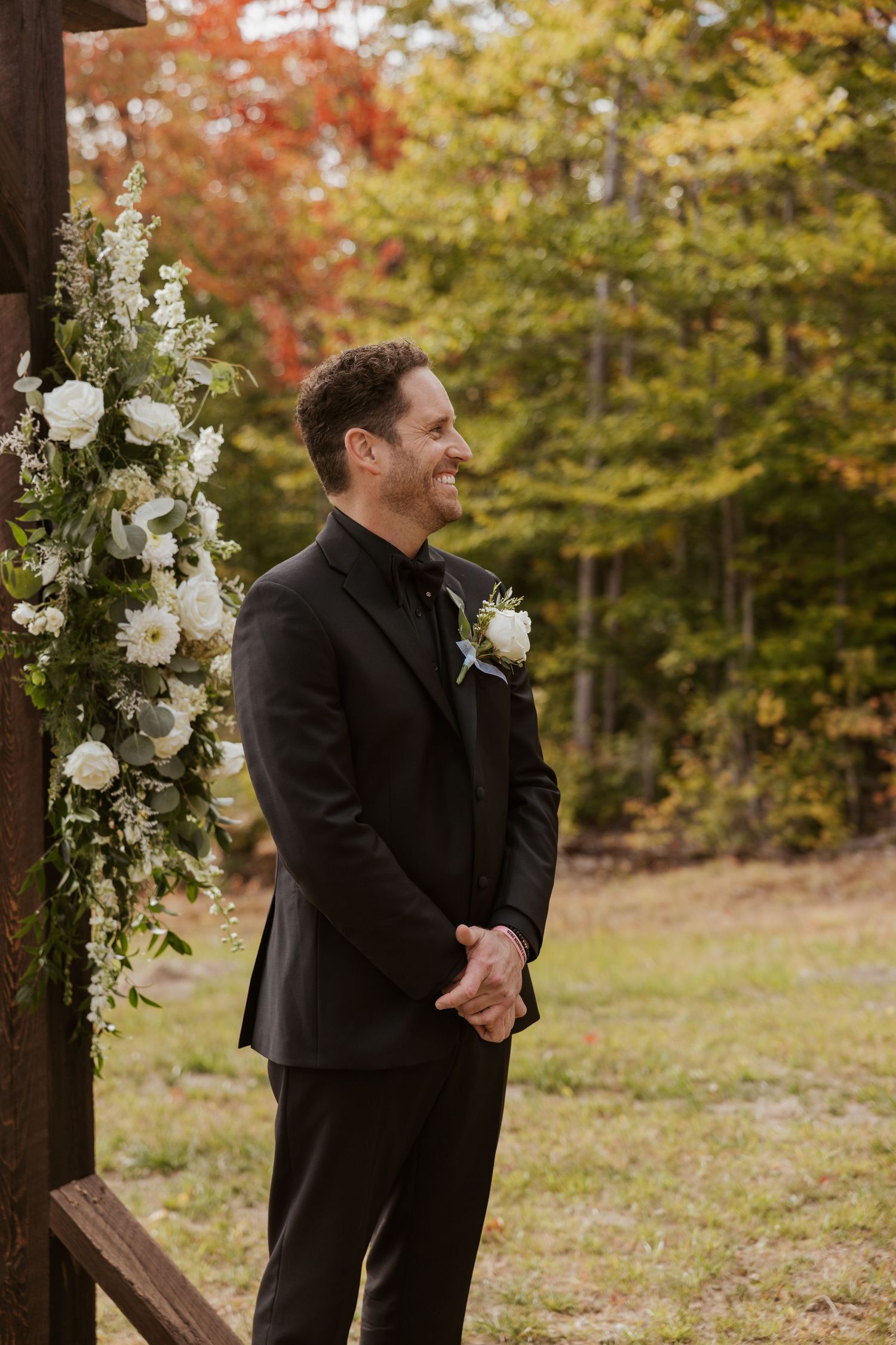 Smiling groom in black suit with white boutonniere stands at floral arch during outdoor fall wedding ceremony.
