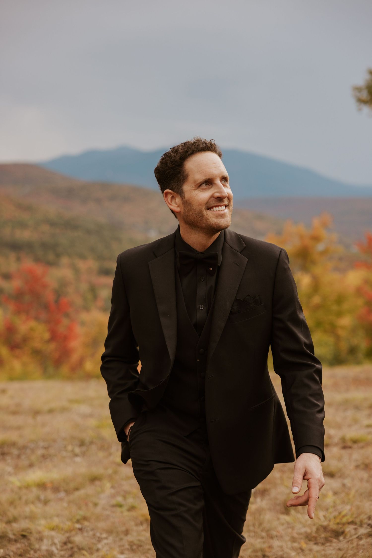 Groom in black tuxedo smiling against autumn mountain landscape with colorful fall foliage.