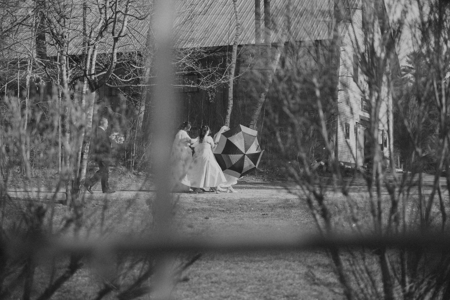 Black and white photo of people in white dresses holding umbrellas outdoors near a barn.