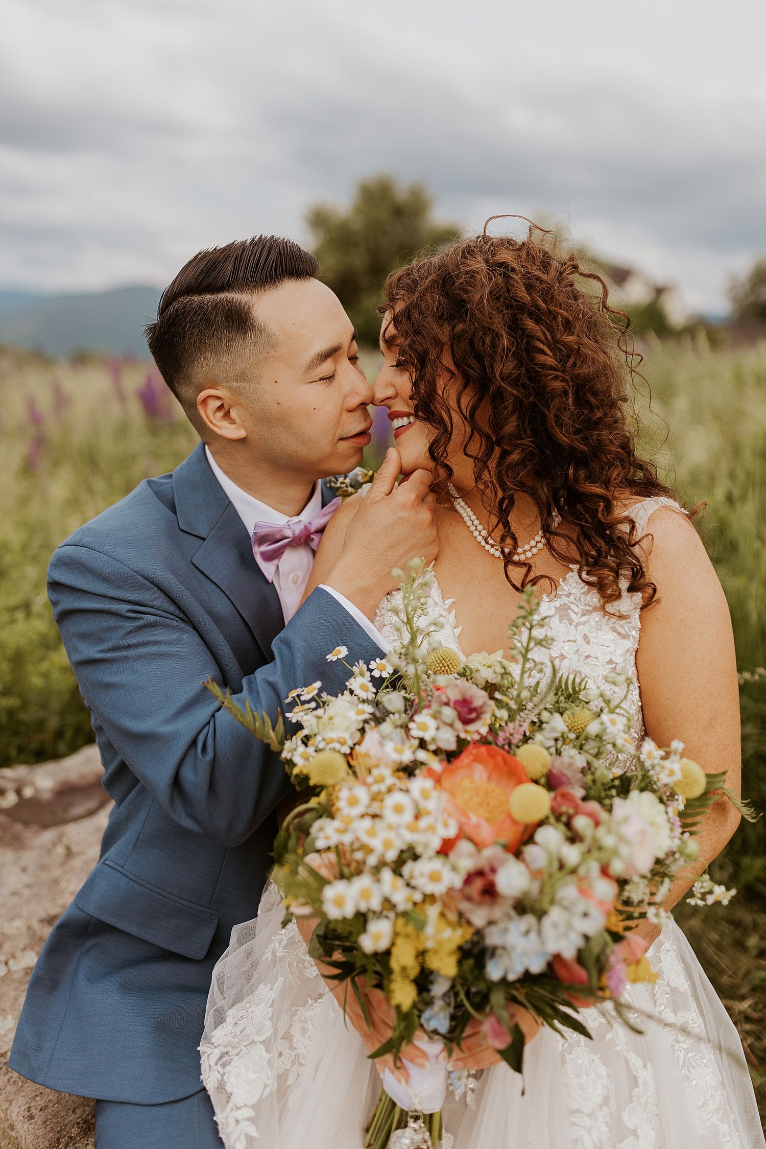 Groom in blue suit with pink bow tie tenderly touches brides face as she holds colorful wildflower bouquet outdoors.