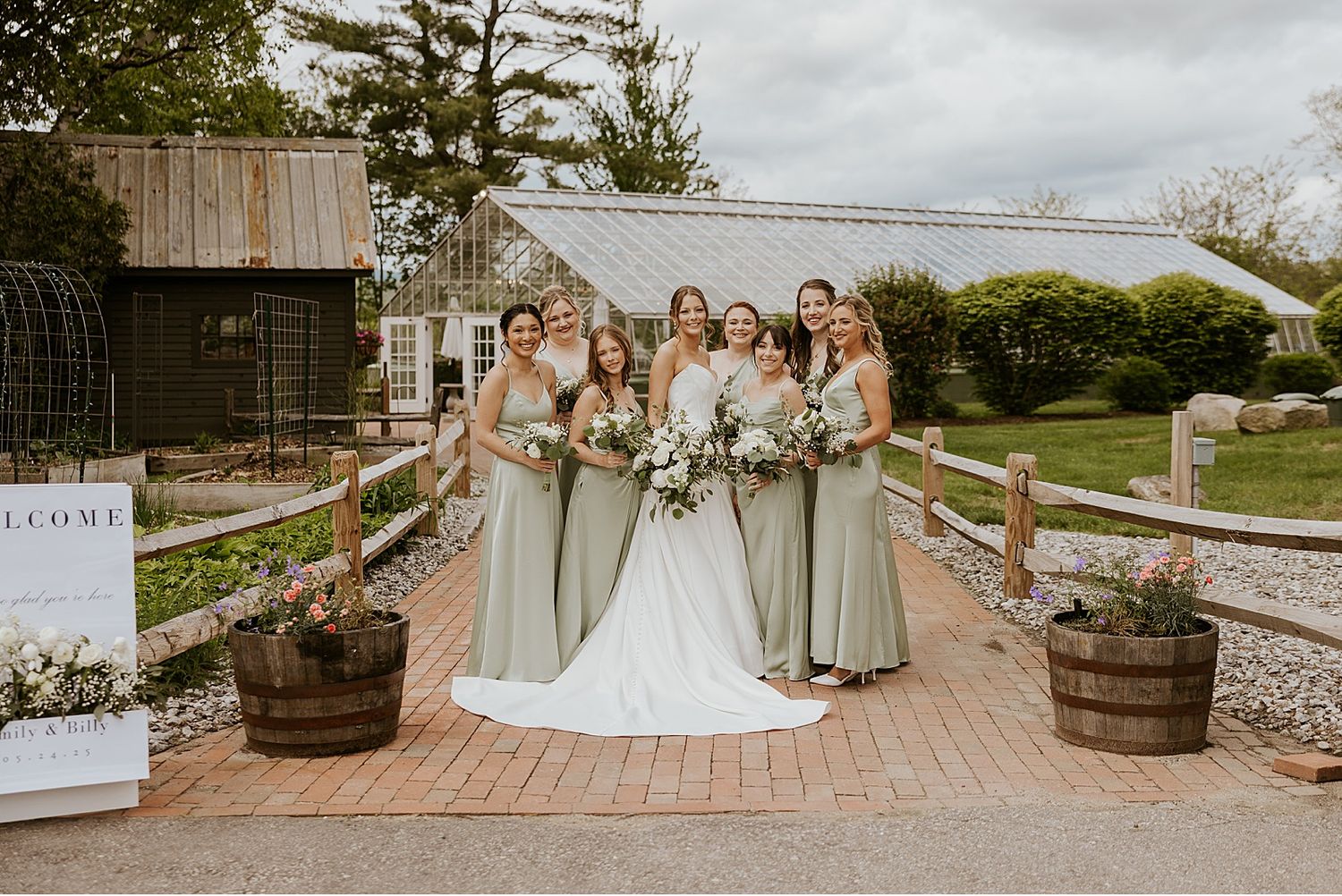 Wedding party poses together outdoors at a rustic venue with wooden barrels and brick pathways.
