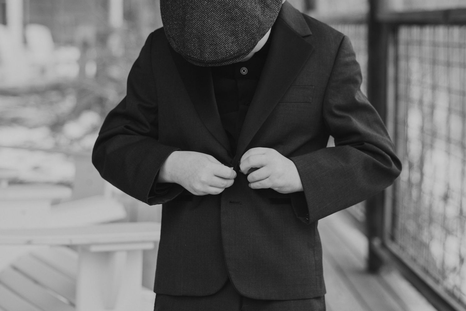 Black and white photo of a young boy in a suit and flat cap buttoning his jacket outdoors.