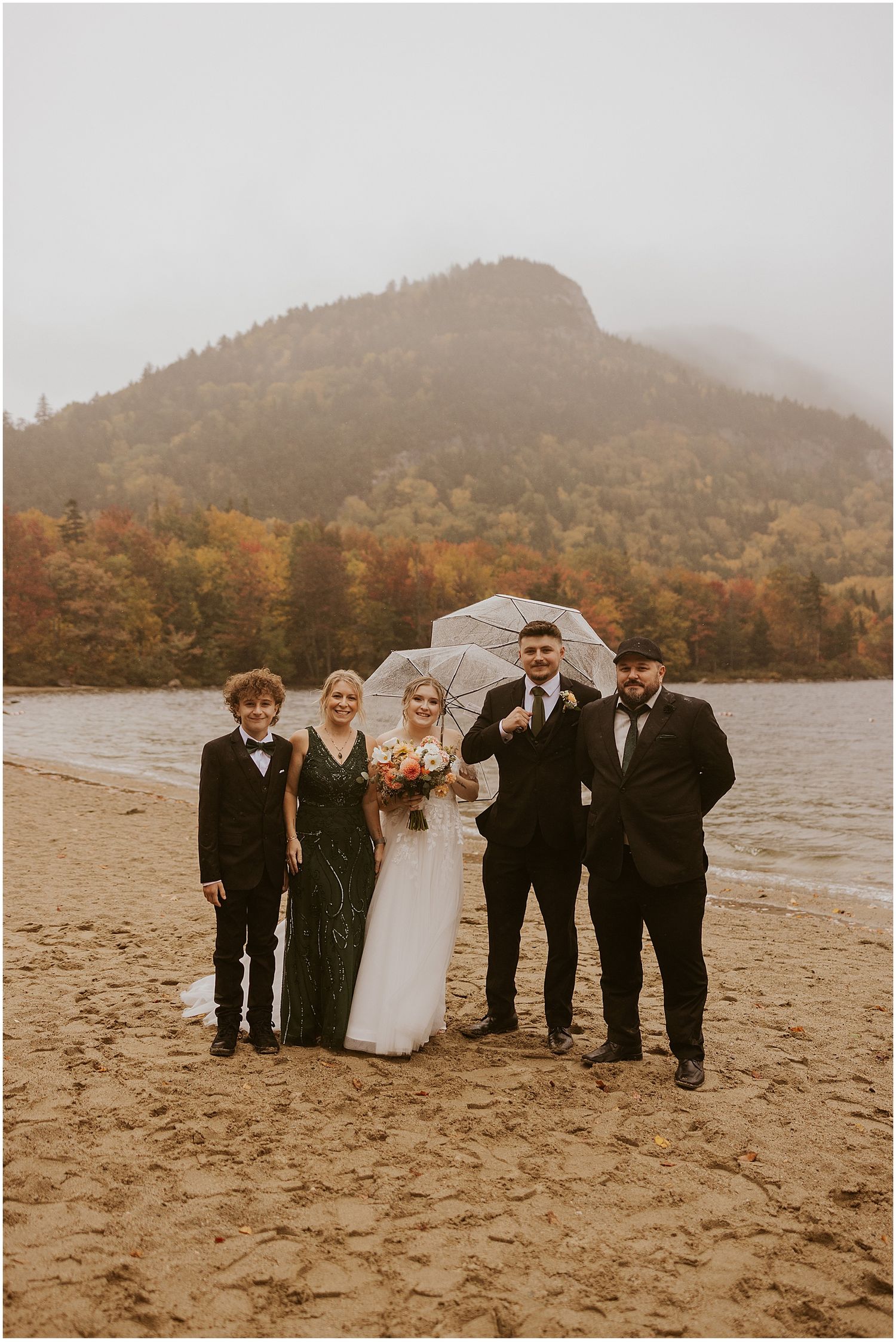 Wedding group photo on a beach with umbrellas against autumn mountain landscape.