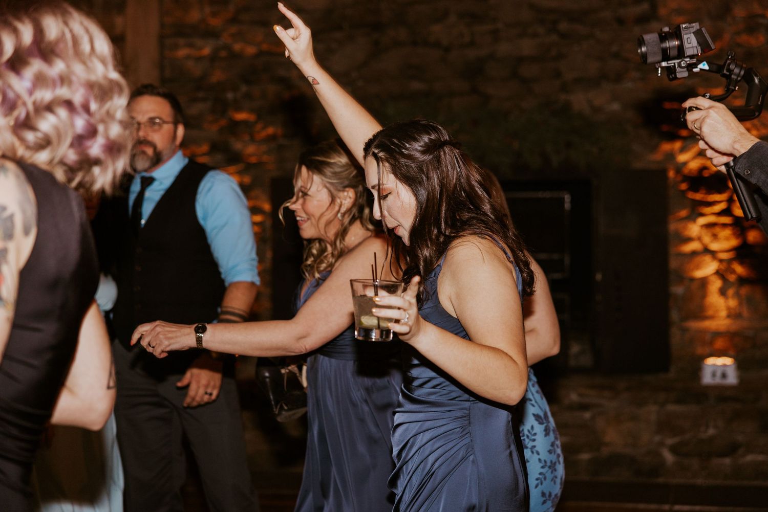 Women in blue bridesmaid dresses dancing joyfully at an indoor wedding reception.