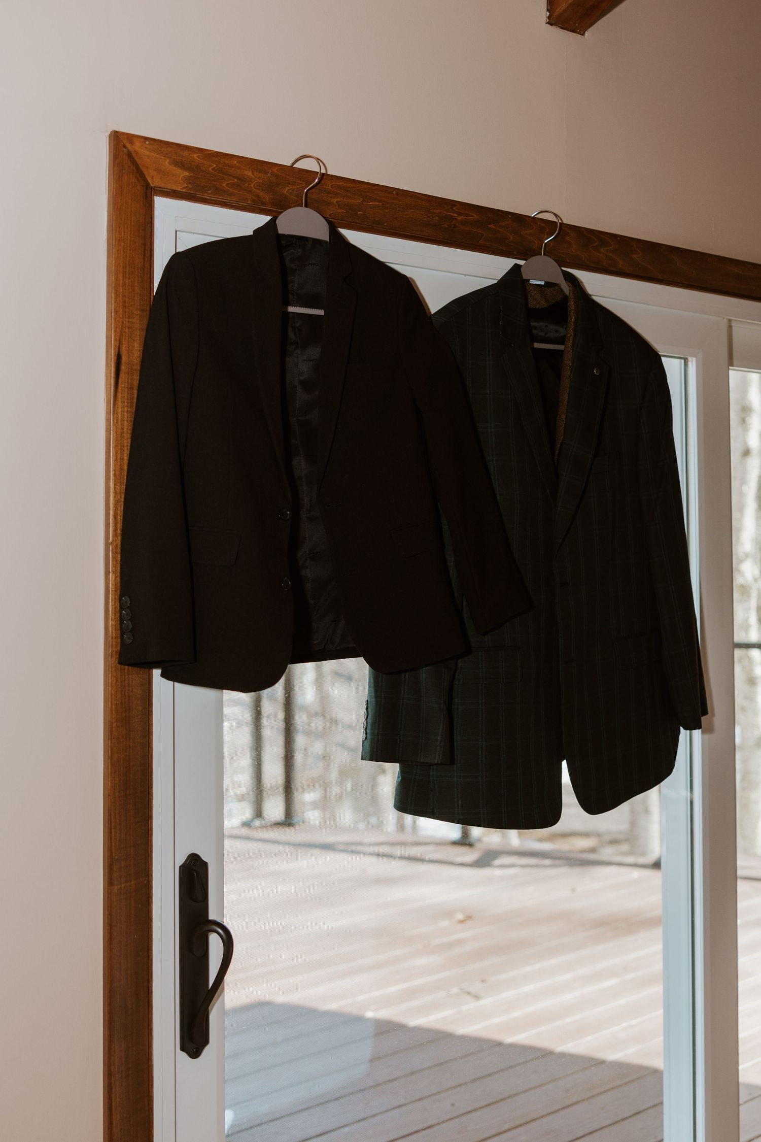 Black suit jackets hang on wooden hangers in a row against a white wall with natural lighting coming through a window.