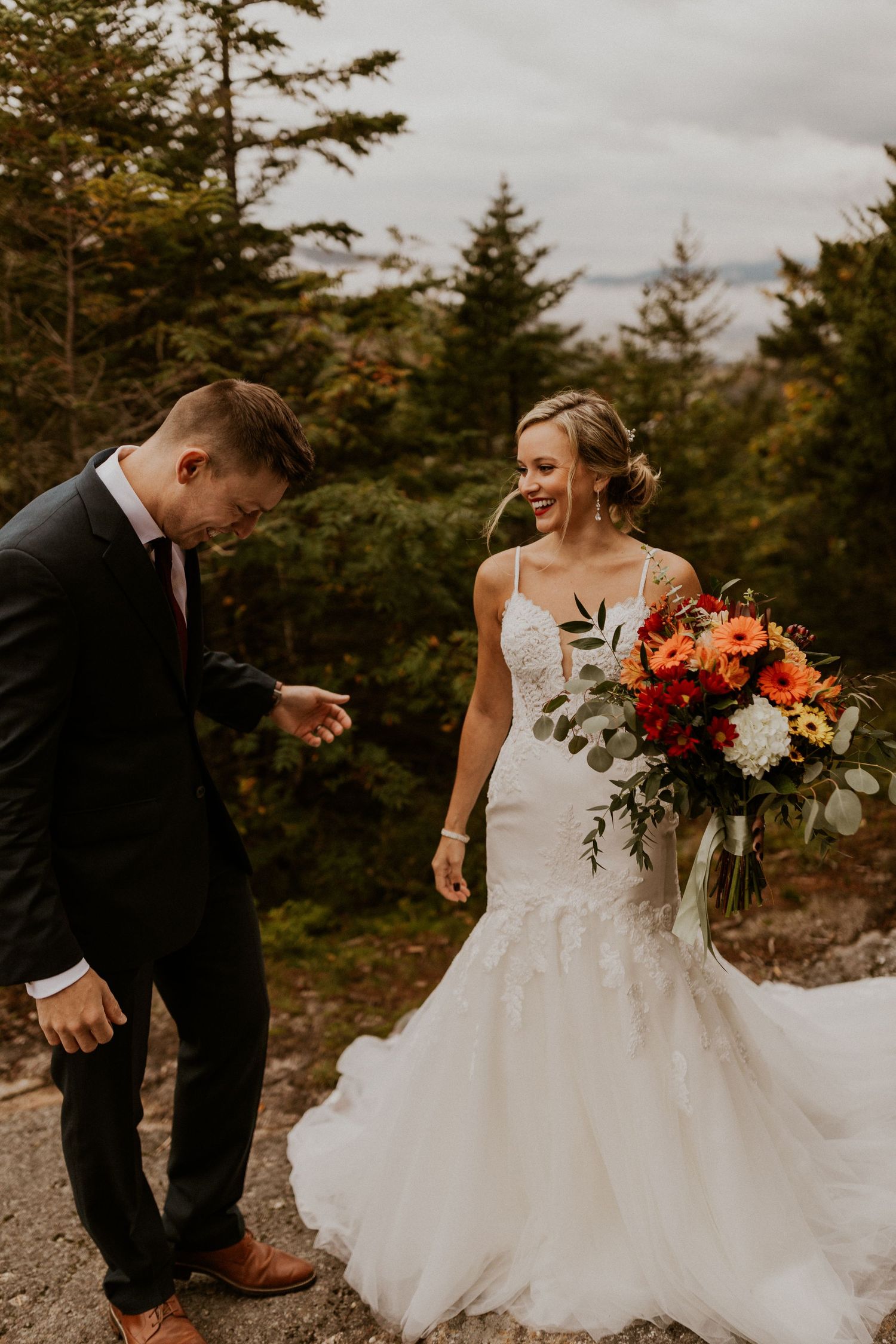 A couple in wedding attire stands on a rocky mountain overlook with pine trees in the background.