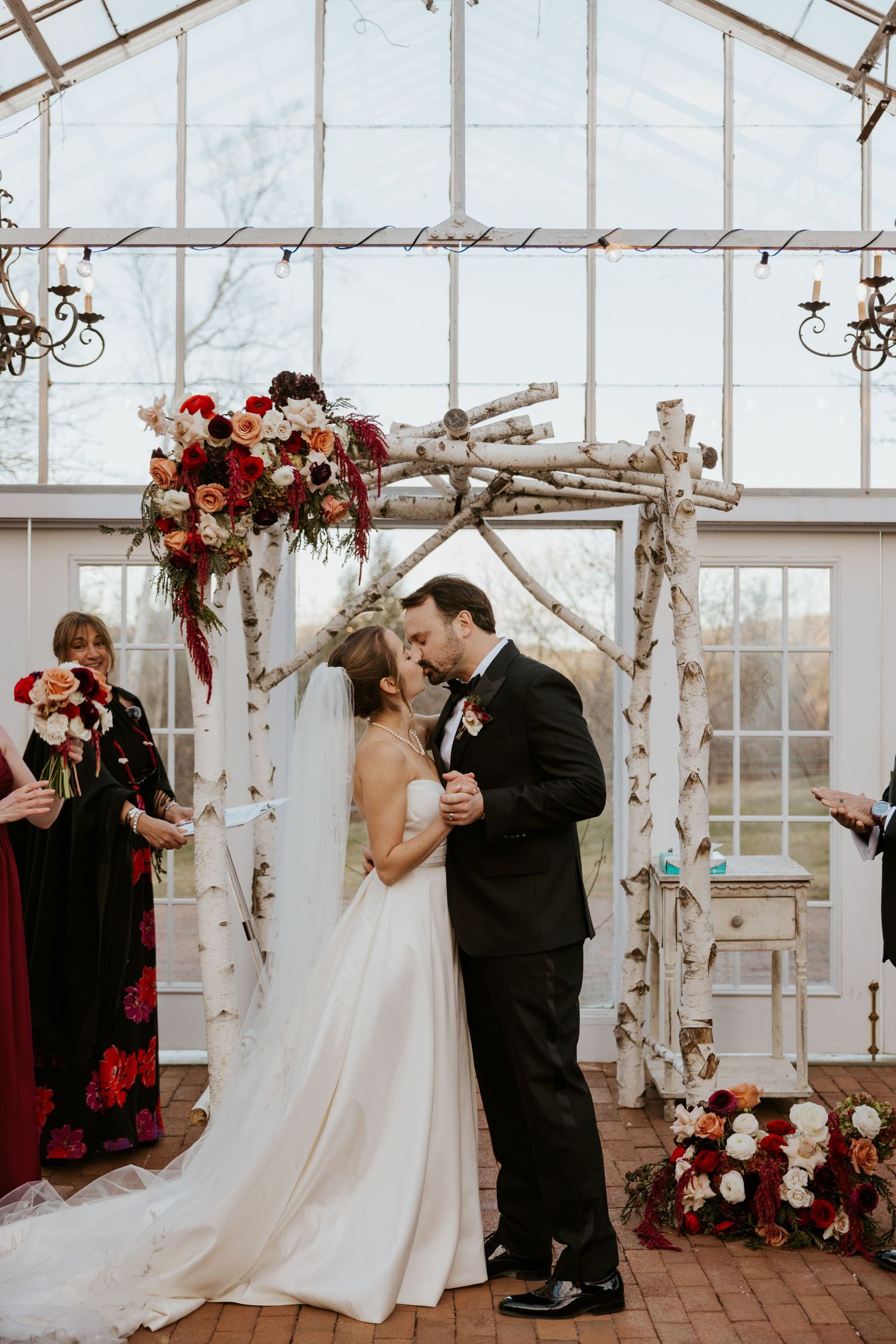 Bride and groom share first kiss under a rustic birch arch adorned with red and white floral arrangements.