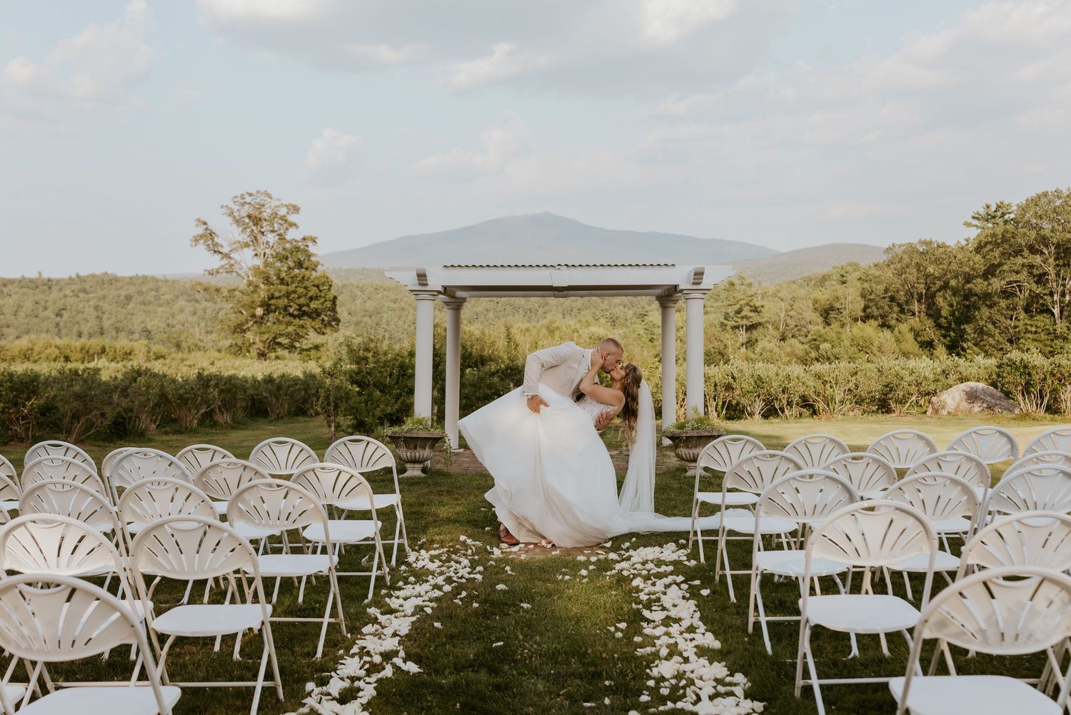 Bride and groom sharing romantic dip kiss at end of white chair lined aisle with mountain backdrop.