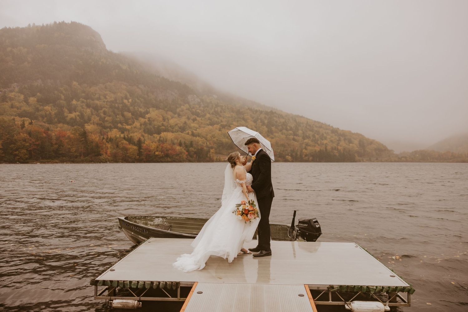 Bride and groom kiss on a lake dock in the rain surrounded by vibrant autumn mountain scenery.