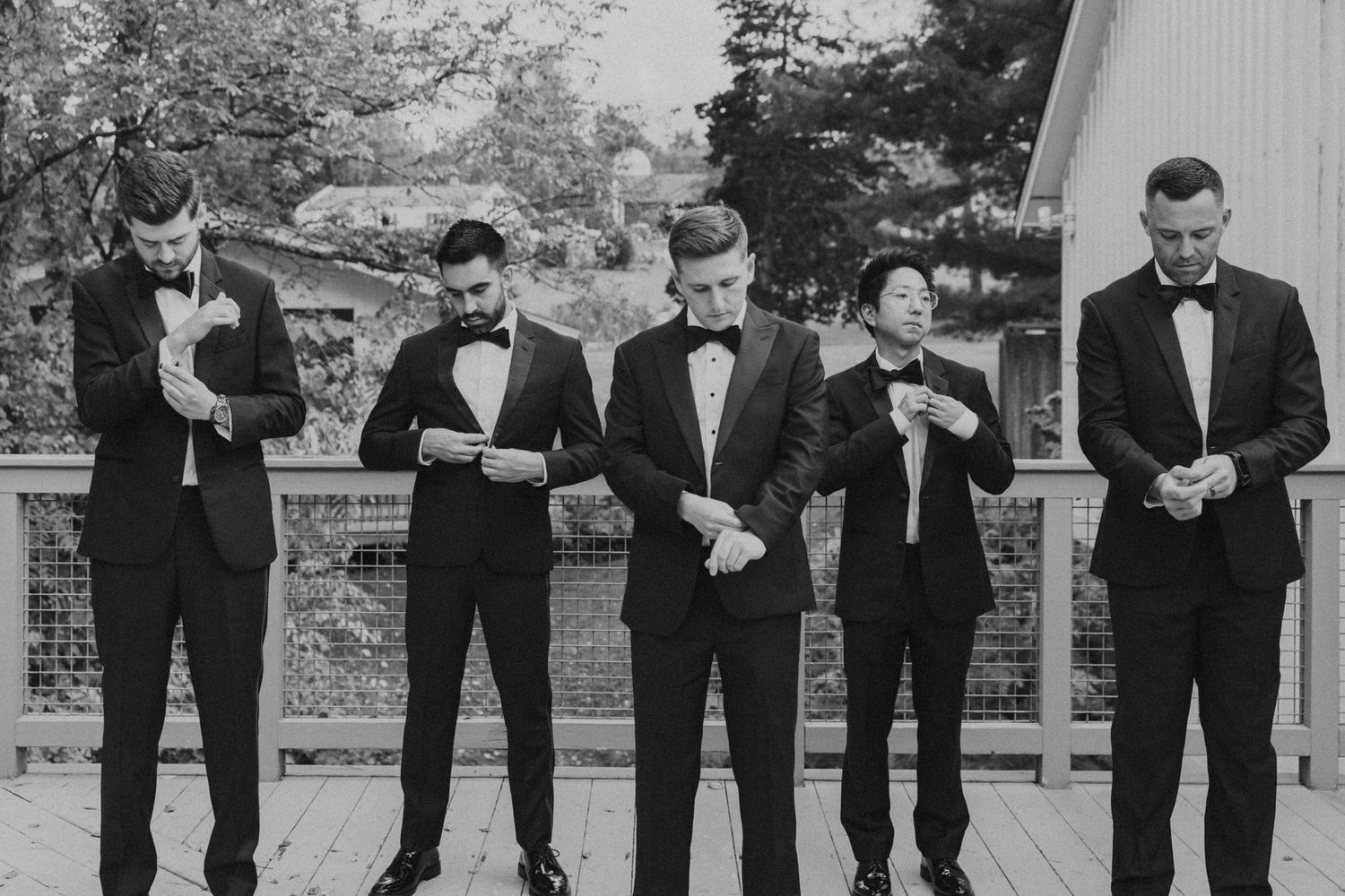 Black and white photo of five groomsmen in tuxedos adjusting cufflinks on an outdoor deck.