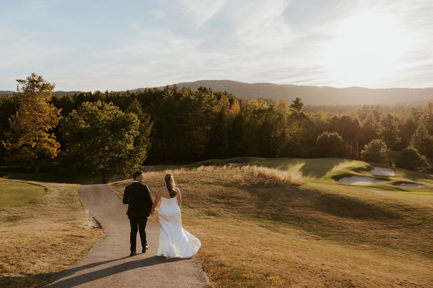 Bride and groom walk along winding path through golden autumn golf course at sunset.
