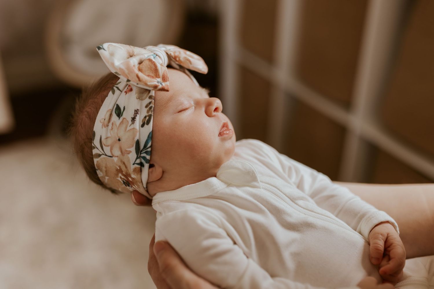 Close-up of a peaceful sleeping newborn baby girl wearing a floral bow headband in a white onesie.