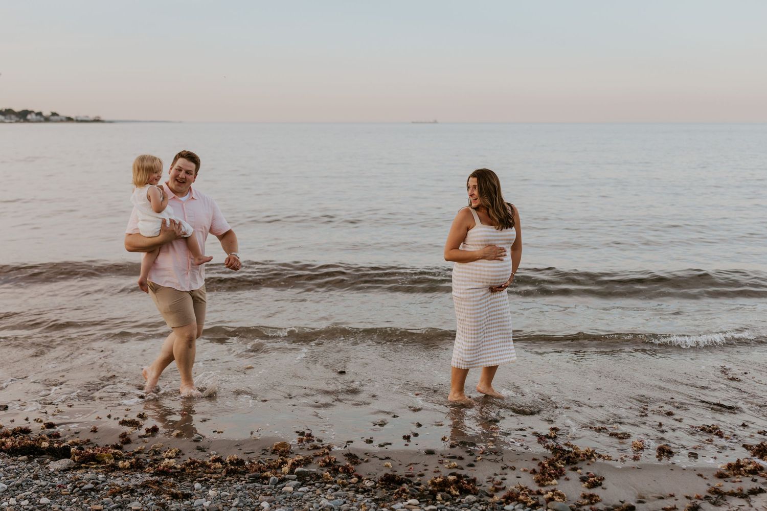 Family maternity photo with father carrying toddler wading in ocean while pregnant mother watches.
