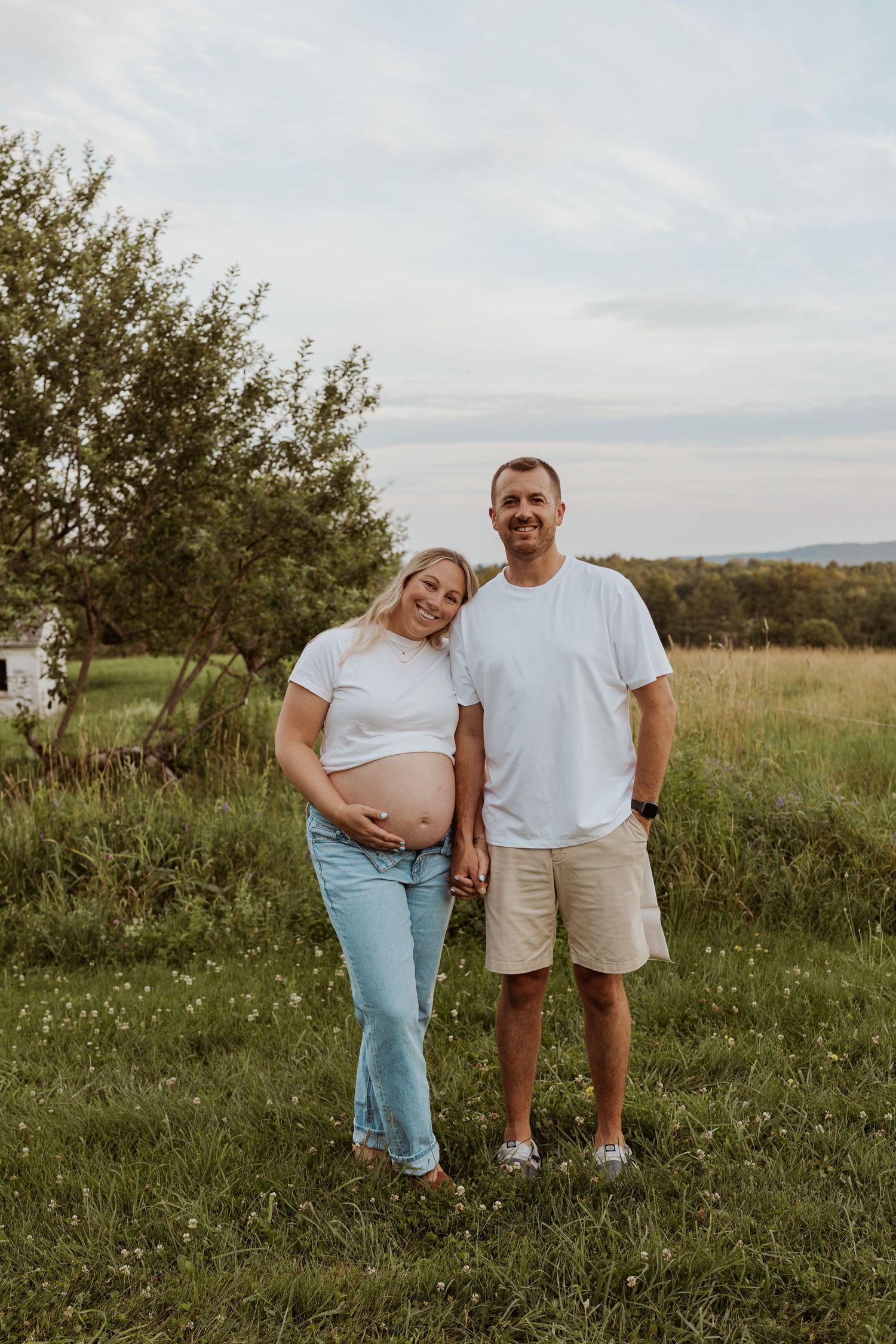 Smiling pregnant couple holding hands in a green field, woman cradling her baby bump in a white tee and jeans.