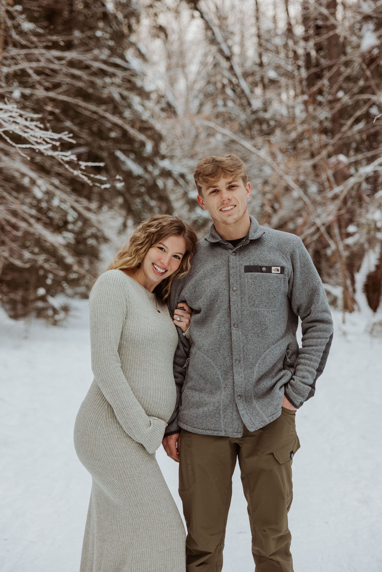 Smiling pregnant couple poses together in snowy winter forest, woman in fitted gray knit dress.