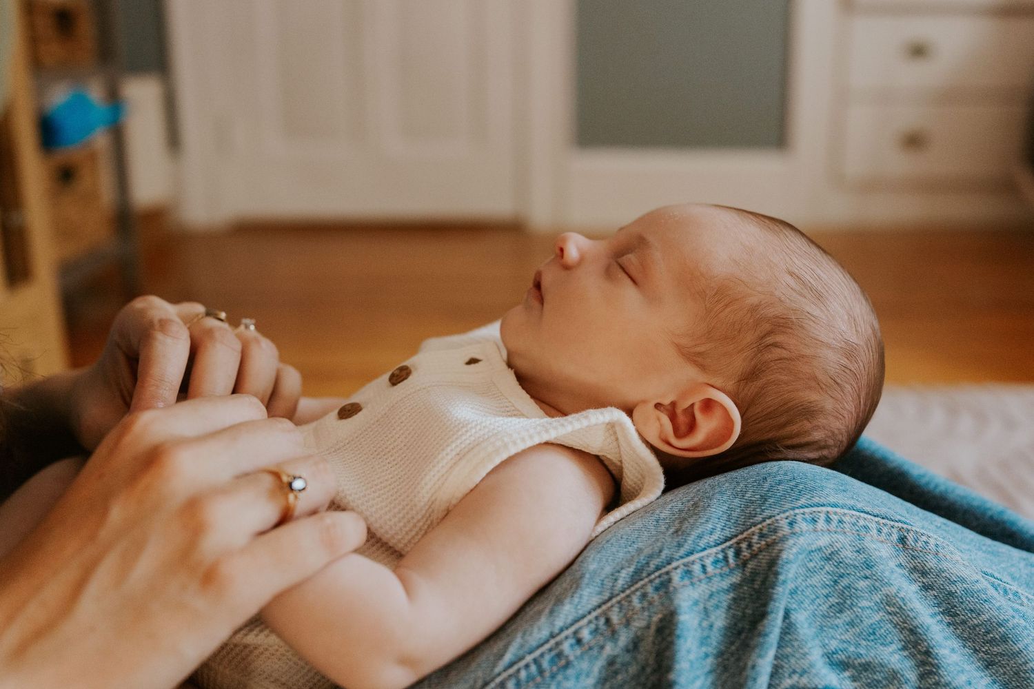 Peaceful sleeping newborn in cream onesie cradled on parent's lap with gentle natural lighting.